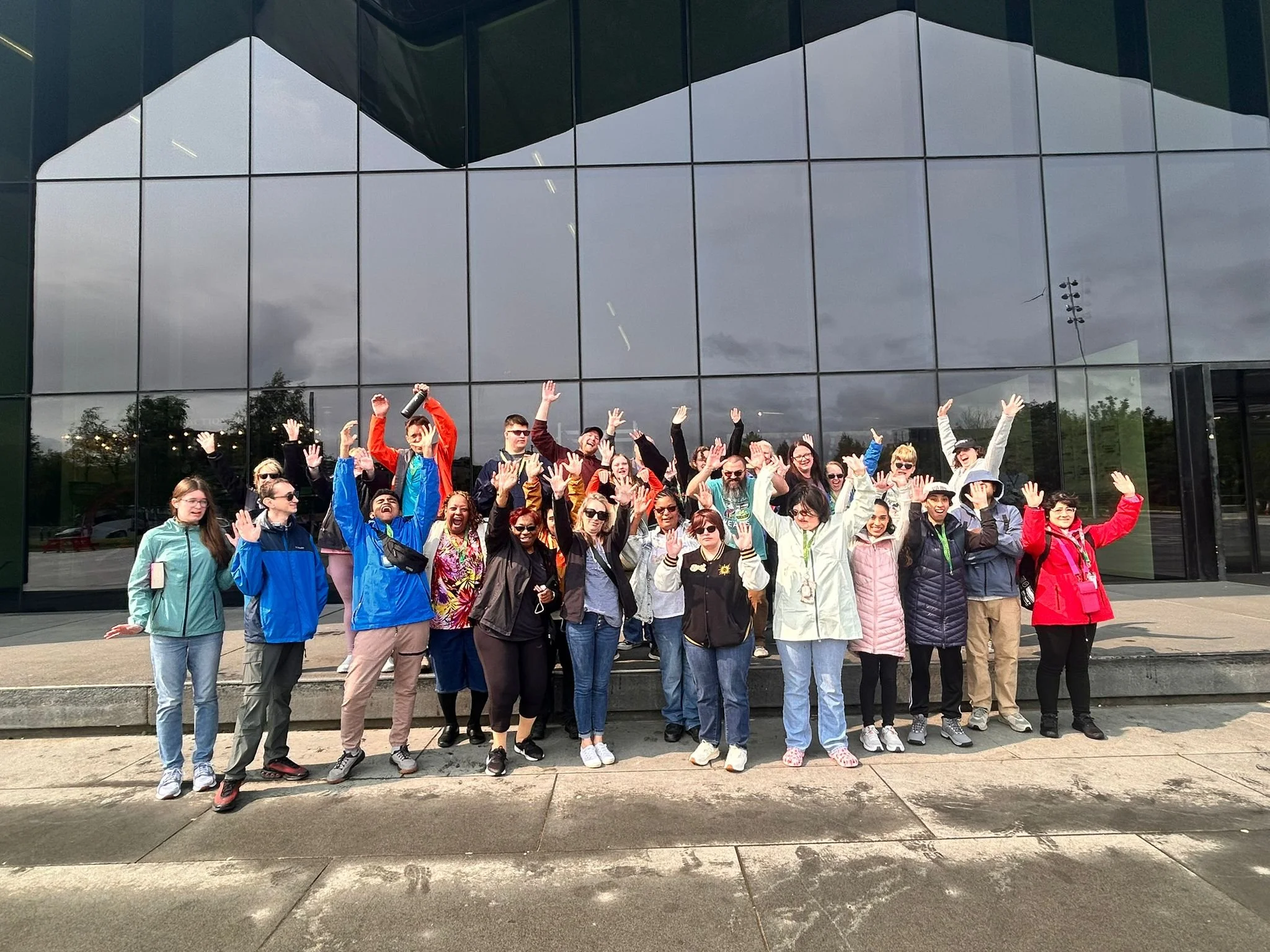 Large group of individuals with a disability jump for joy in front of a big glass window of a museum in Scotland.
