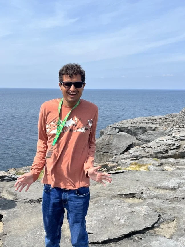 A student in a salmon-colored long sleeve shirt with a sunflower lanyard. He is smiling haply at the camera, behind him there are many rocks that break to a vast ocean behind the smiling traveler. This is just outside the Cliffs of Moher in Ireland. 