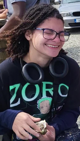Camera captures a young lady with braided hair and glasses smiling wiht her eyes closed and looking to the right of the pitcutre. She has an oyster in her hands.