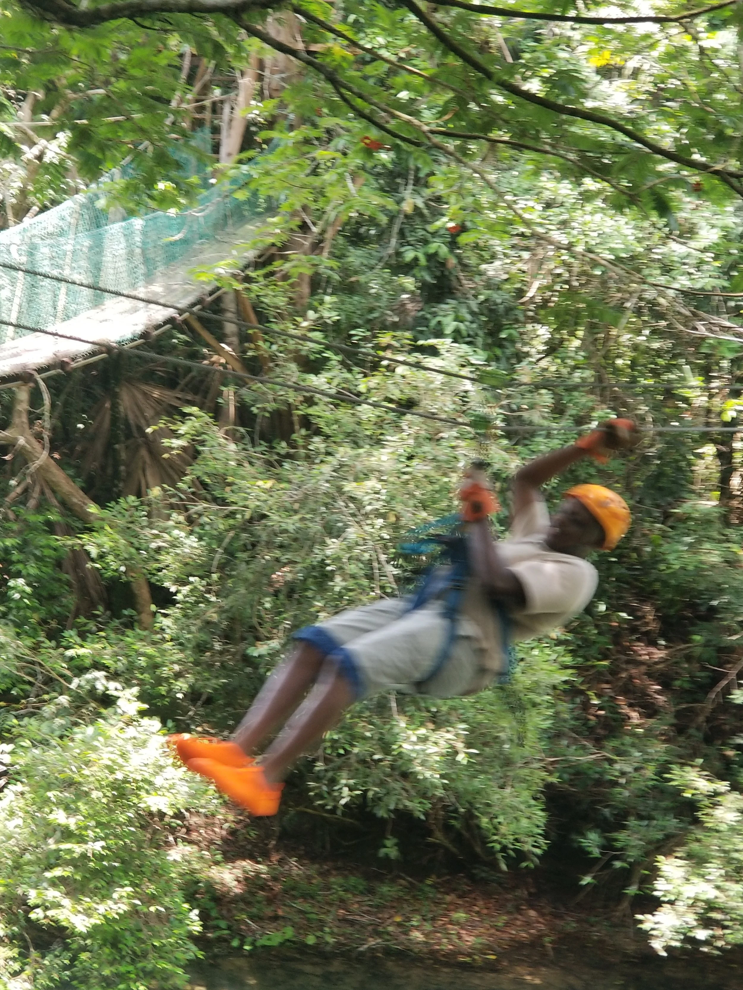 A young man in the group, wearing orange shoes, white/blue pants, cream shirt and orange helmet and gloves is zip Lining through the Jungle-which is green all around him, with the exception of below him, which has a river. He is blurry, indicating he