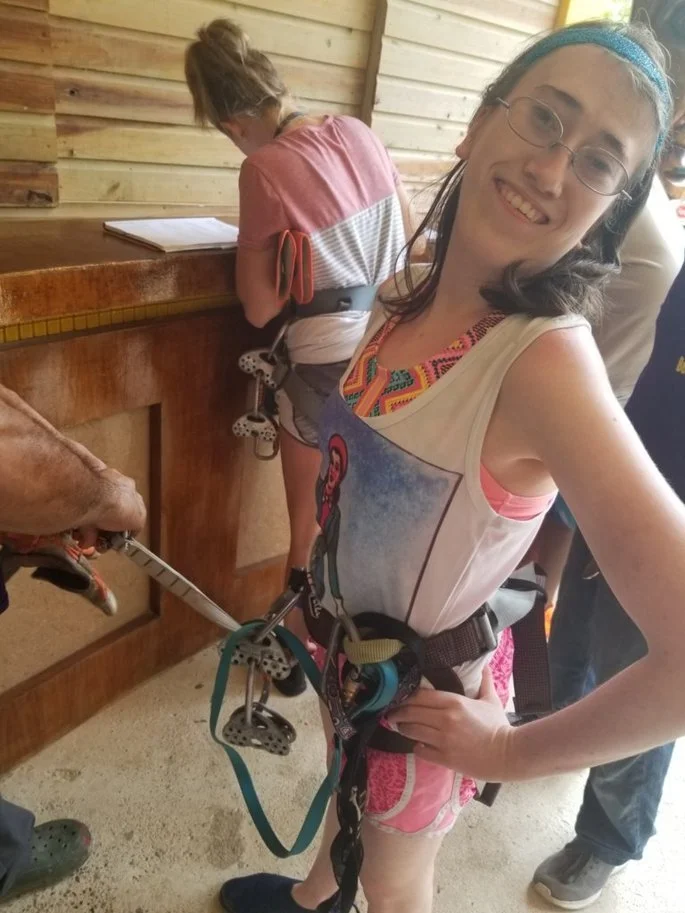 Young woman smiles at the camera with a harness on, getting ready to zip line