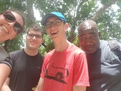 Four people in a photo, smiling toward the camera with trees in the background- waiting for a bus in Belize