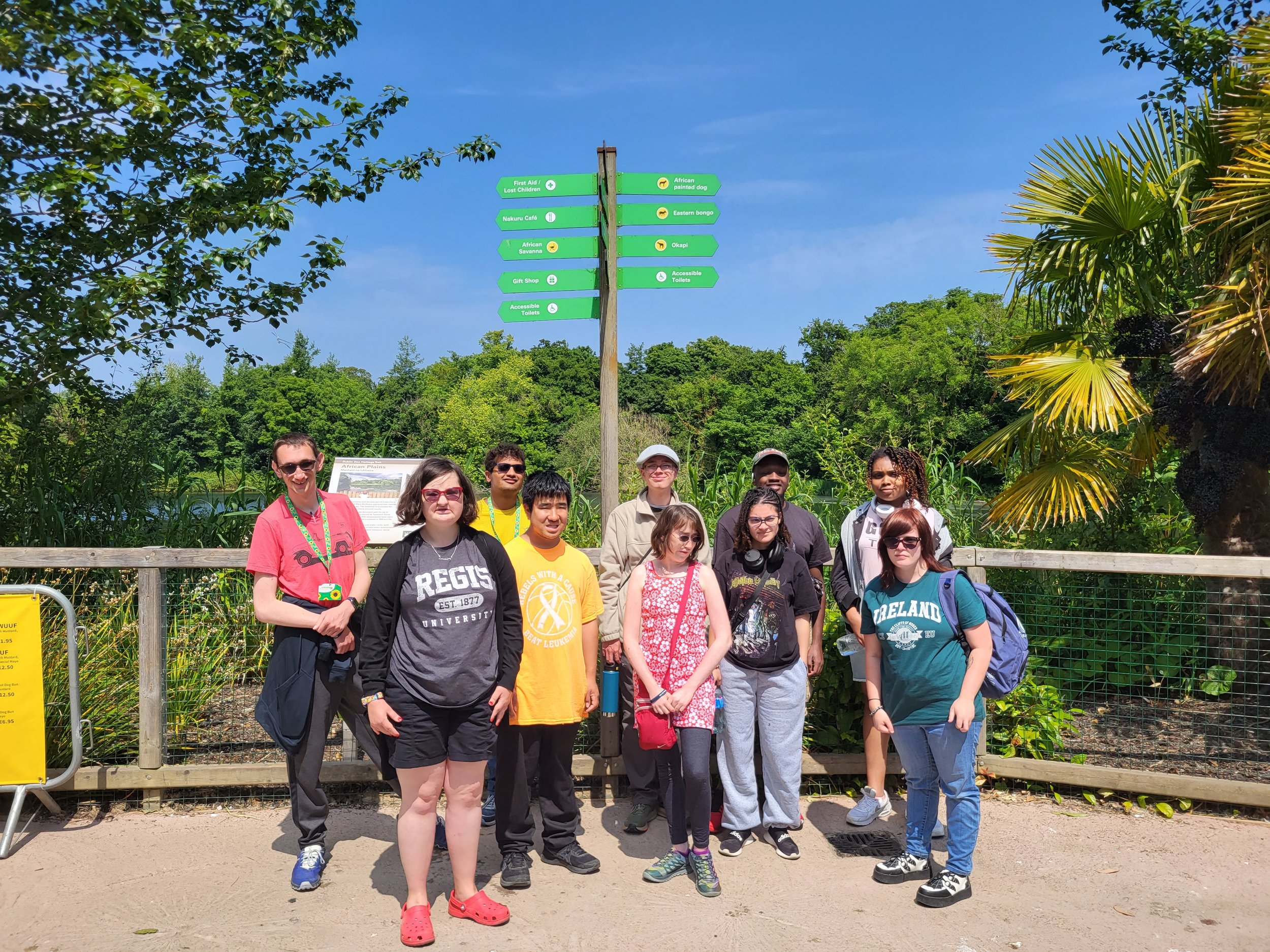 a group of individuals stands in front of a sign and loads of green trees. Everyone is smiling at the camera. Behind them on the sign there are two different arrow directions, indicating where to go to see which animal is where. we are at the Dublin 