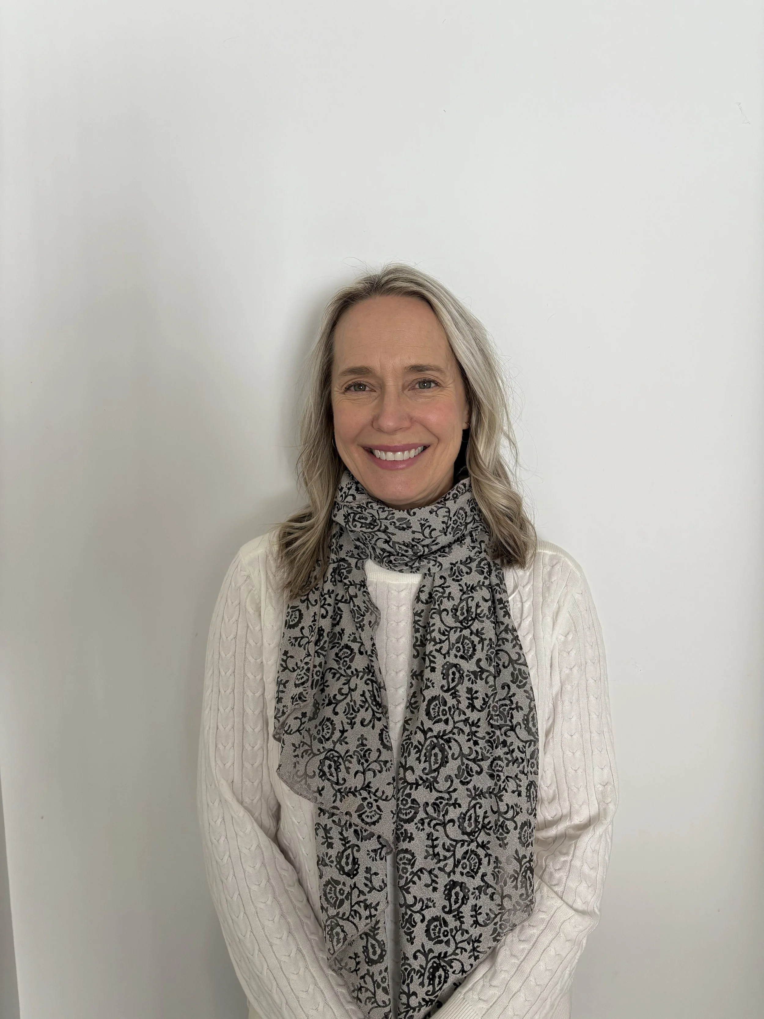 ALT TEXT: Sue stands in front of a white background. She has lighter blonde hair that goes to her shoulders. She is smiling at the camera wearing a white long sleeve white sweater and a black patterned scarf.