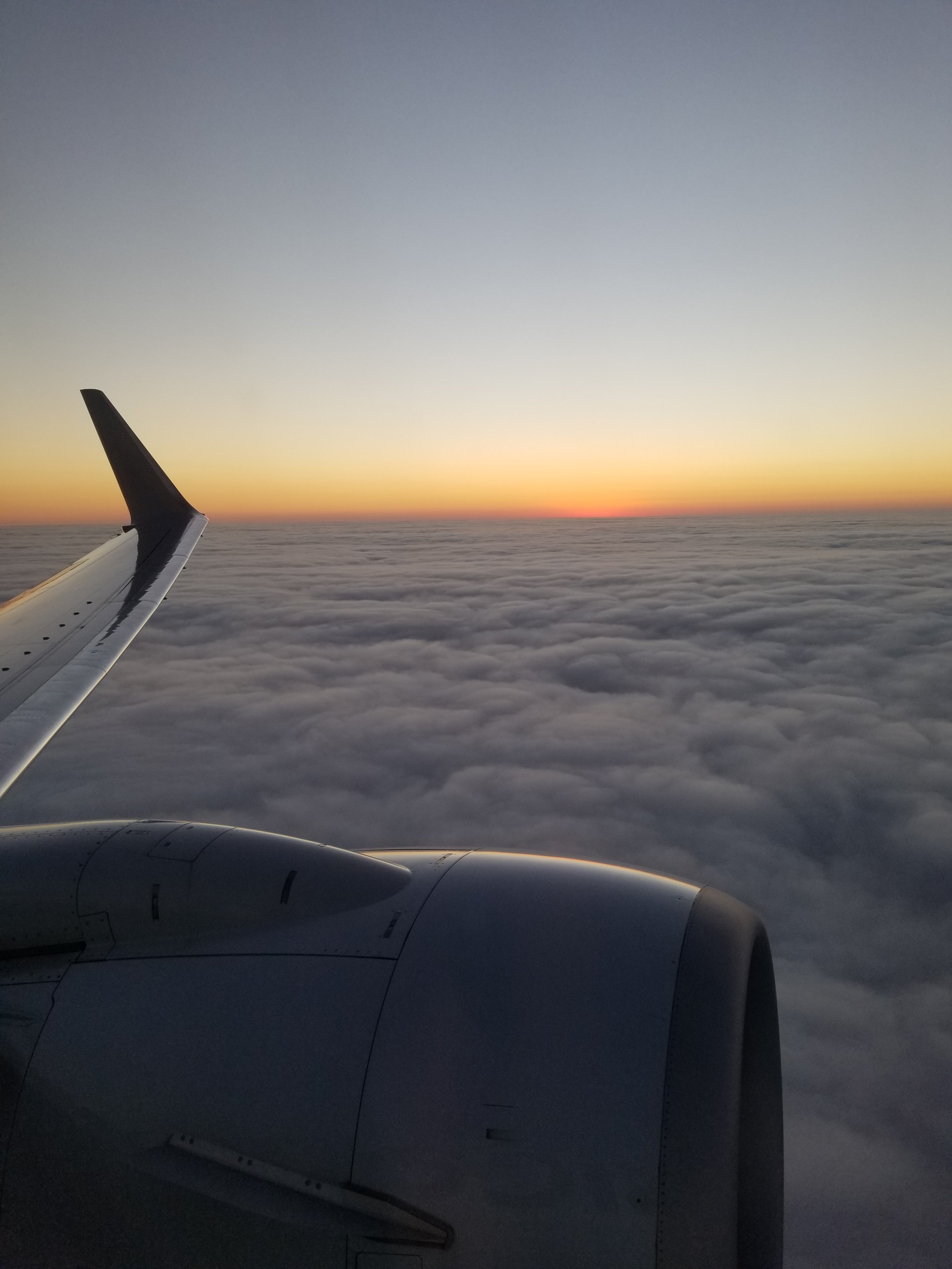 picture from the window of a plane. you can see the wing on the left whith engine below. in the distance there are clouds are far as the eye can see, it is sunset and sky furthest away is made up of orange and yellow hues.