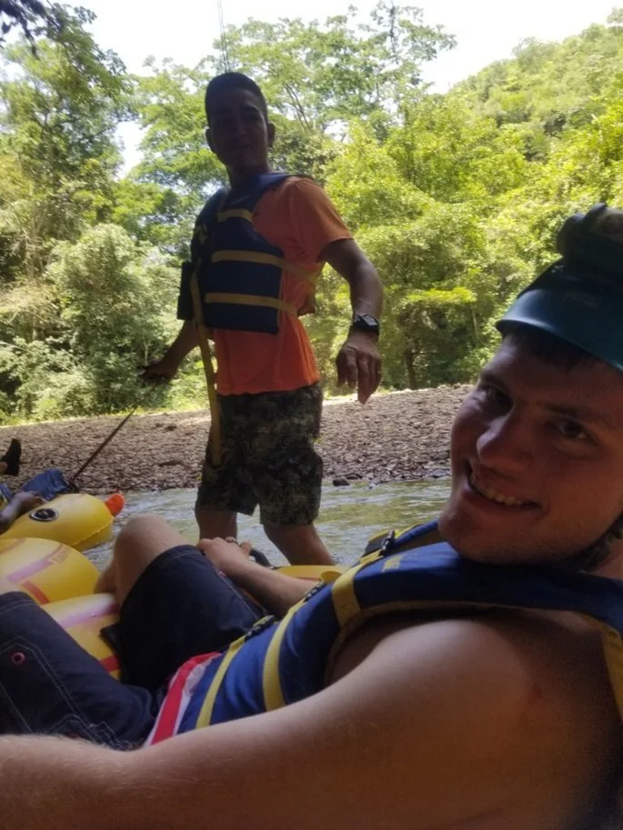 Two men smile at the camera, one is sitting in a tube with a life vest, another man is standing, guiding the tube through the caves of Belize 