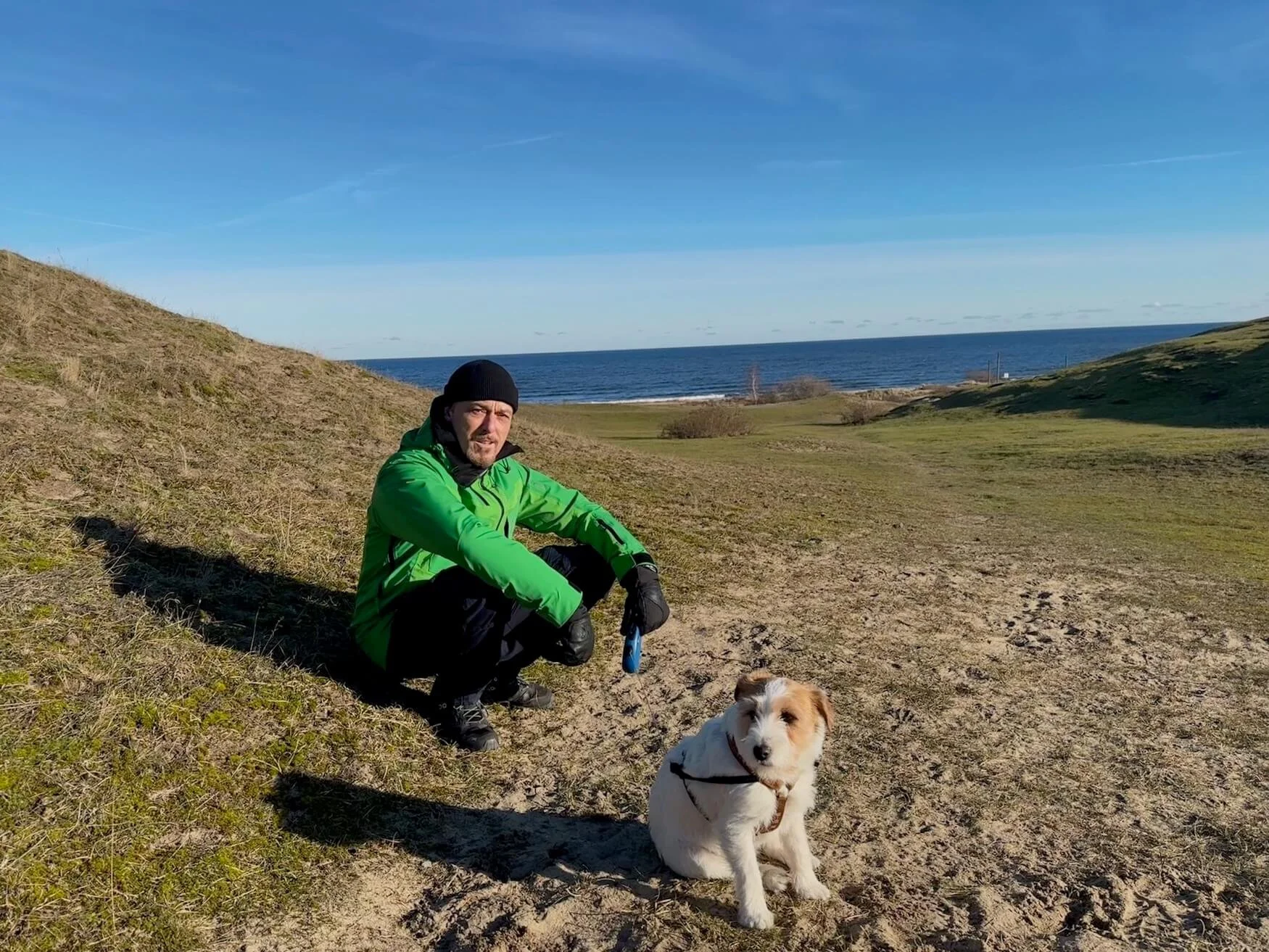 A man in a bright green jacket and black beanie sitting on a grassy and sandy hill near the coast, with a white and brown dog sitting beside him, during a sunny day with a blue sky and ocean in the background.