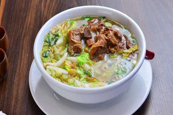 A bowl of beef noodle soup with chopped beef, noodles, leafy greens, green onions, and broth