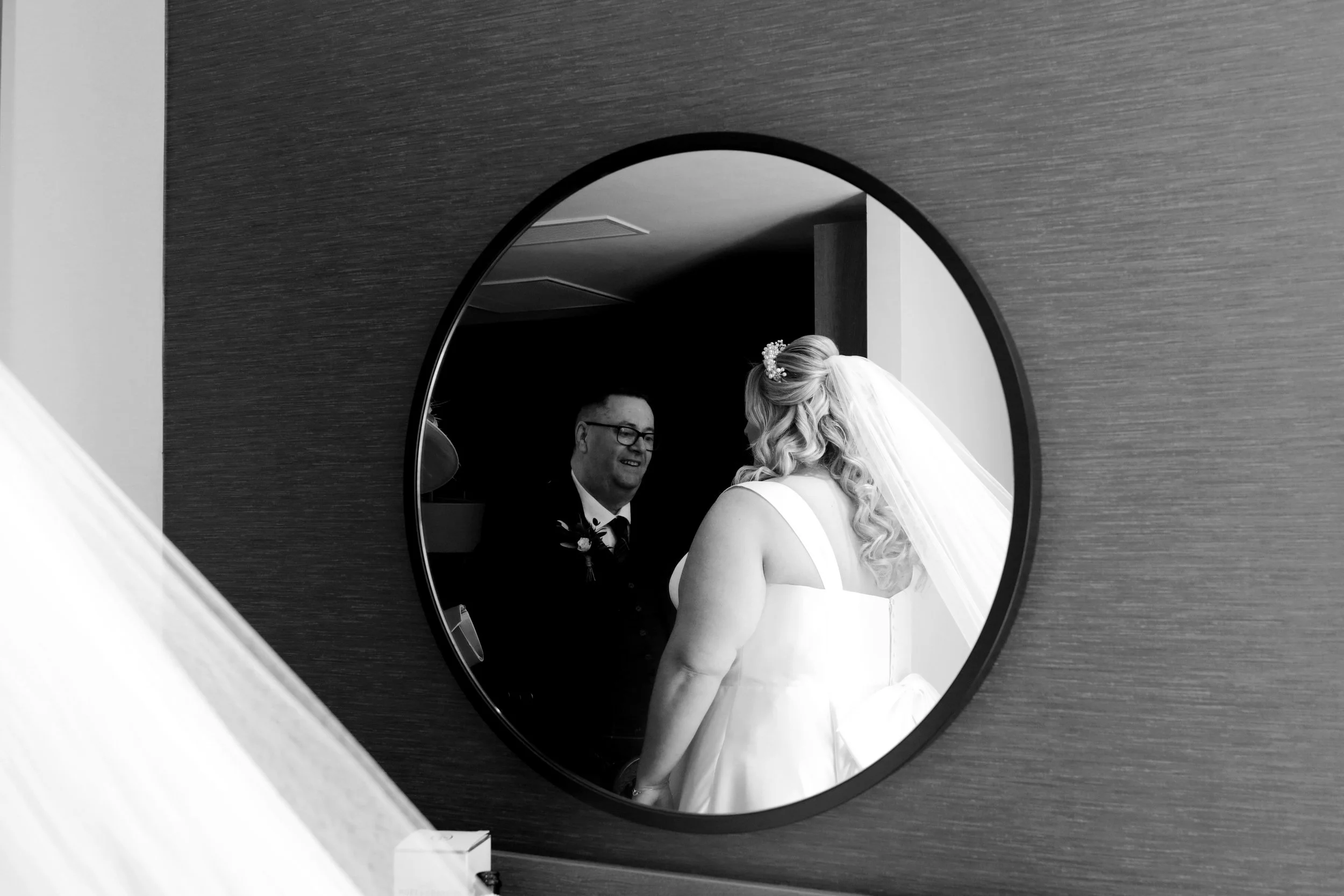 A reflection in a round mirror shows a bride and a groom during a wedding, with the bride in a wedding dress and veil, and the groom in a suit and glasses.