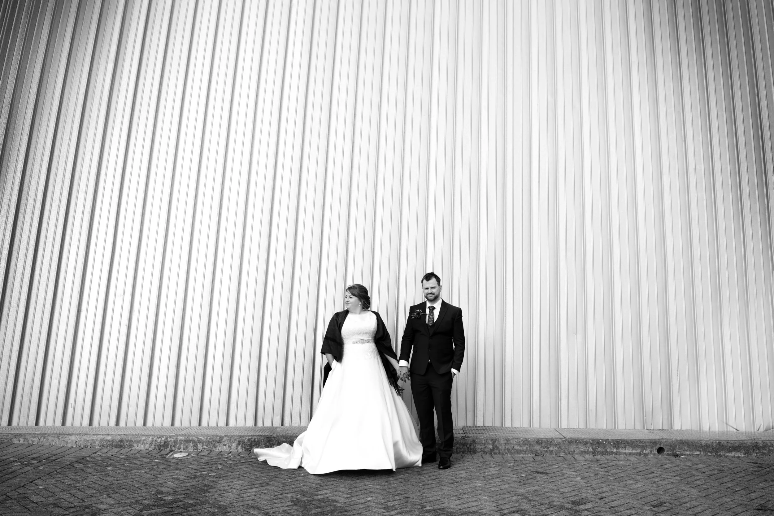 A bride in a white wedding dress and a groom in a black suit holding hands and walking on a brick sidewalk in front of a tall, metallic wall. Glasgow City Centre wedding.Wedding photography Scotland, Wedding Venue Scotland. 