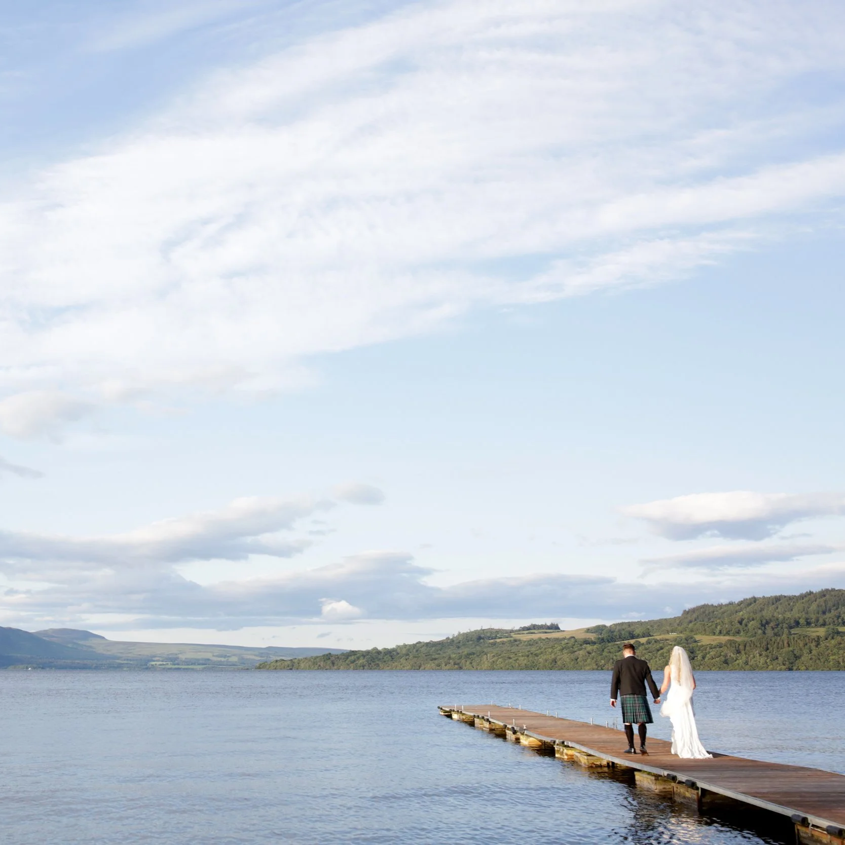 A bride and groom holding hands walking on a wooden pier over a lake with a partly cloudy sky and distant hills.