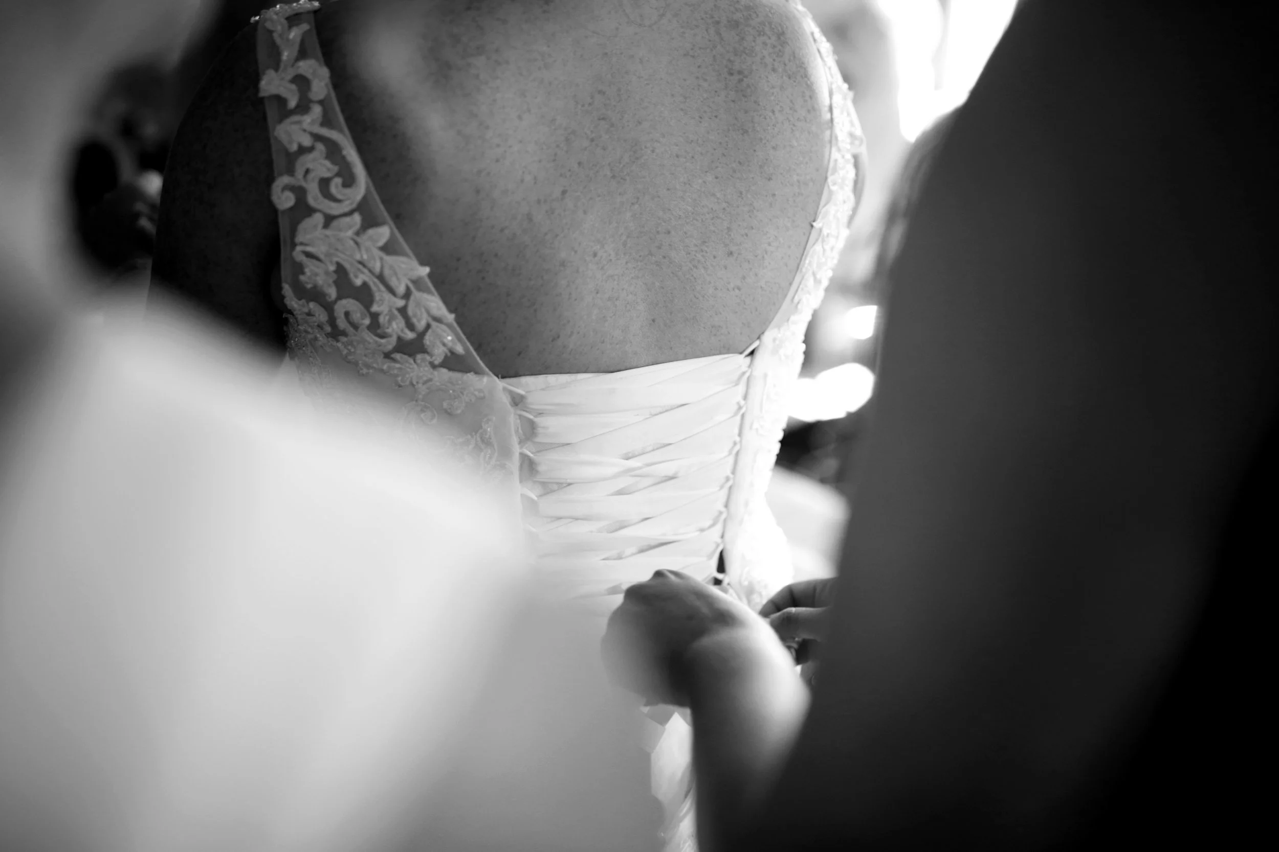 A bride in a wedding dress is being fastened at the back by an assistant, with focus on the bridal gown's laced and ruched details, black and white photo.