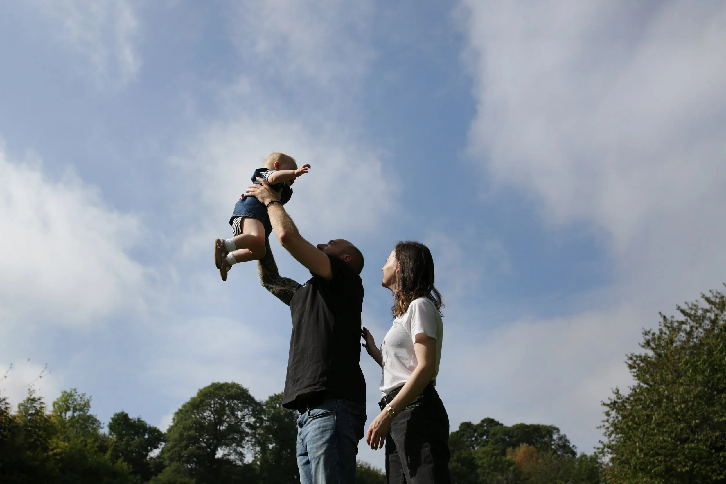 A man lifts a young child into the air while a woman stands nearby, all outdoors under a partly cloudy sky with trees in the background.