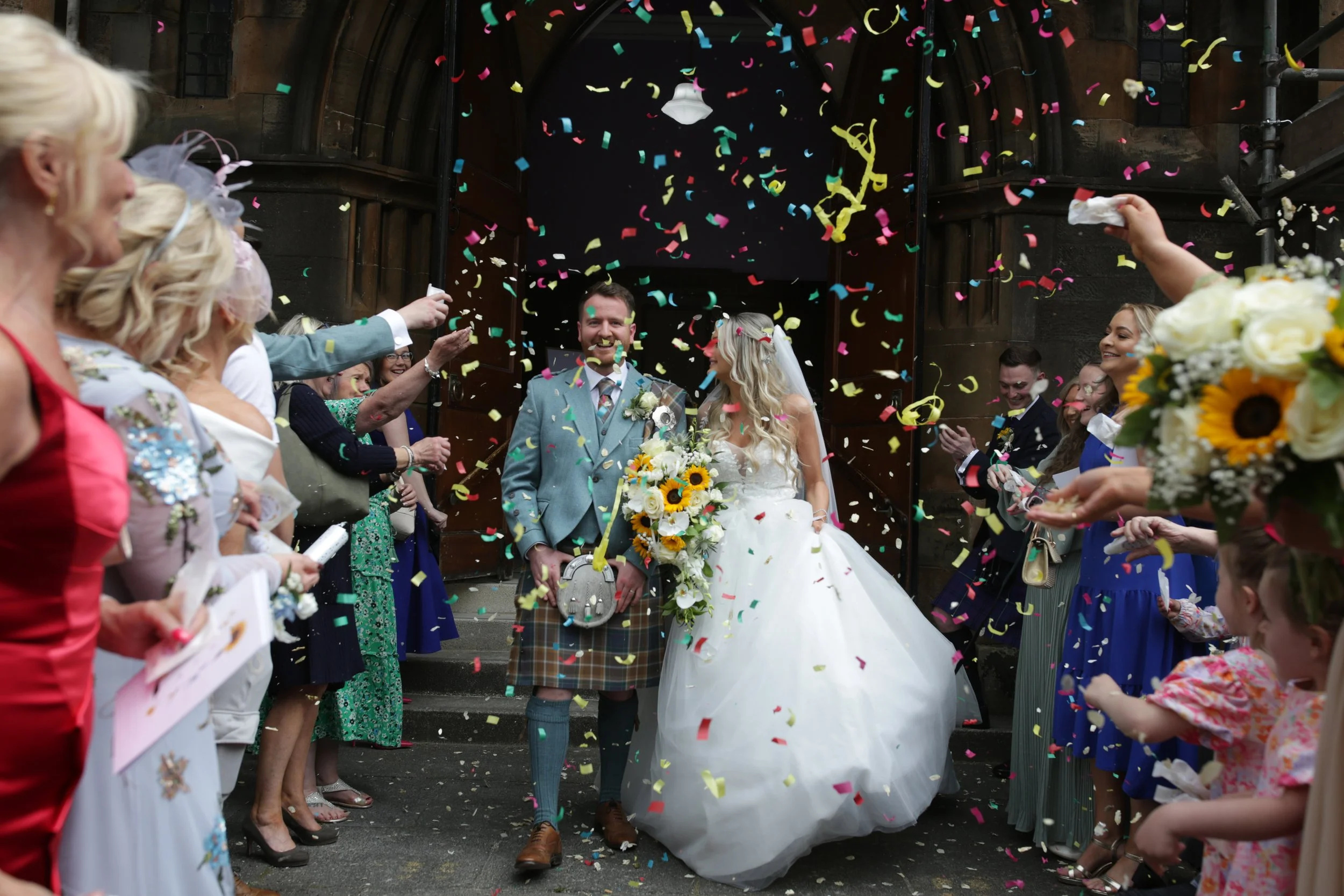 Bride and groom celebrating outside a church with guests throwing colorful confetti.