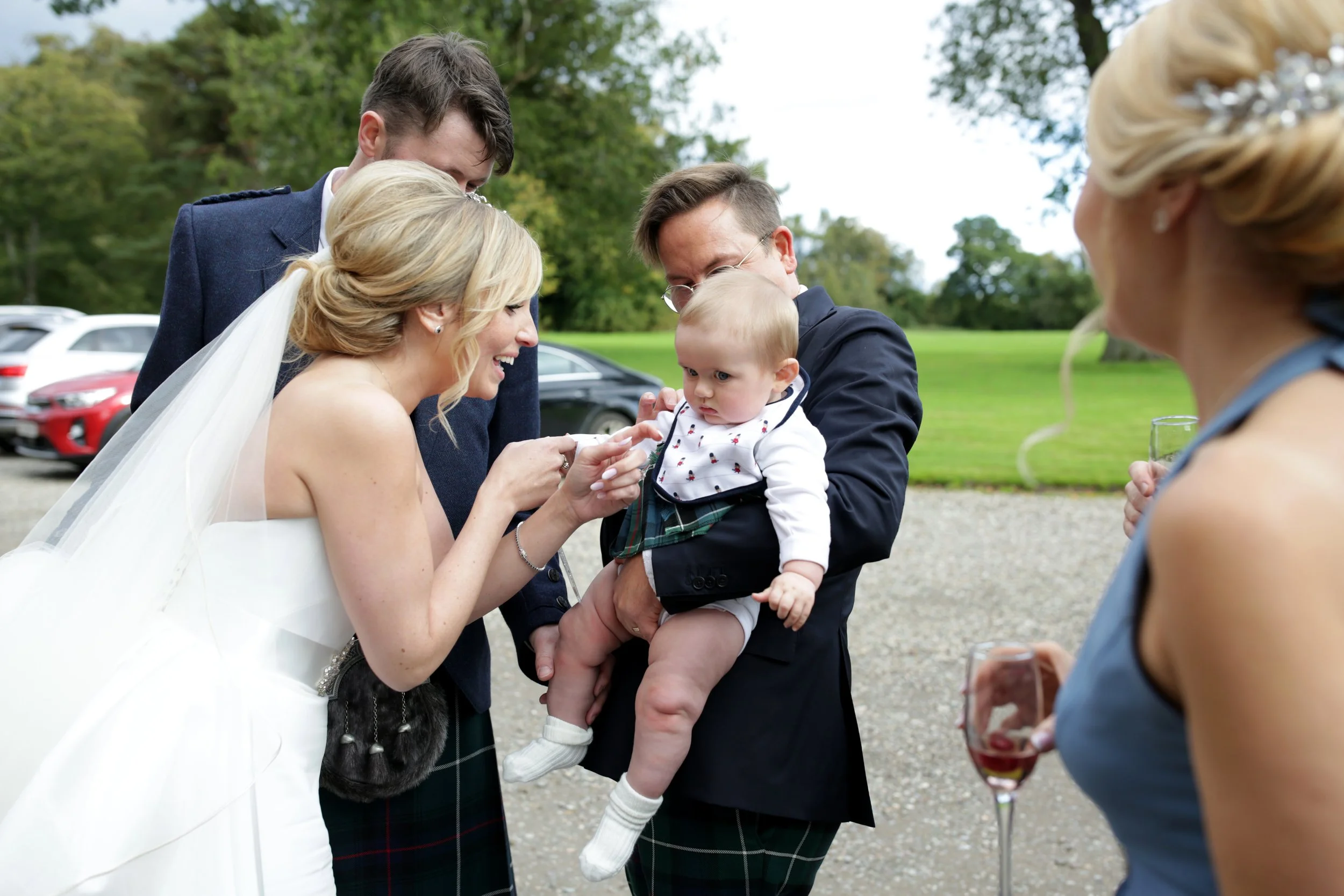 A bride and groom look at a baby held by a man at an outdoor wedding reception, with friends nearby holding drinks and smiling.