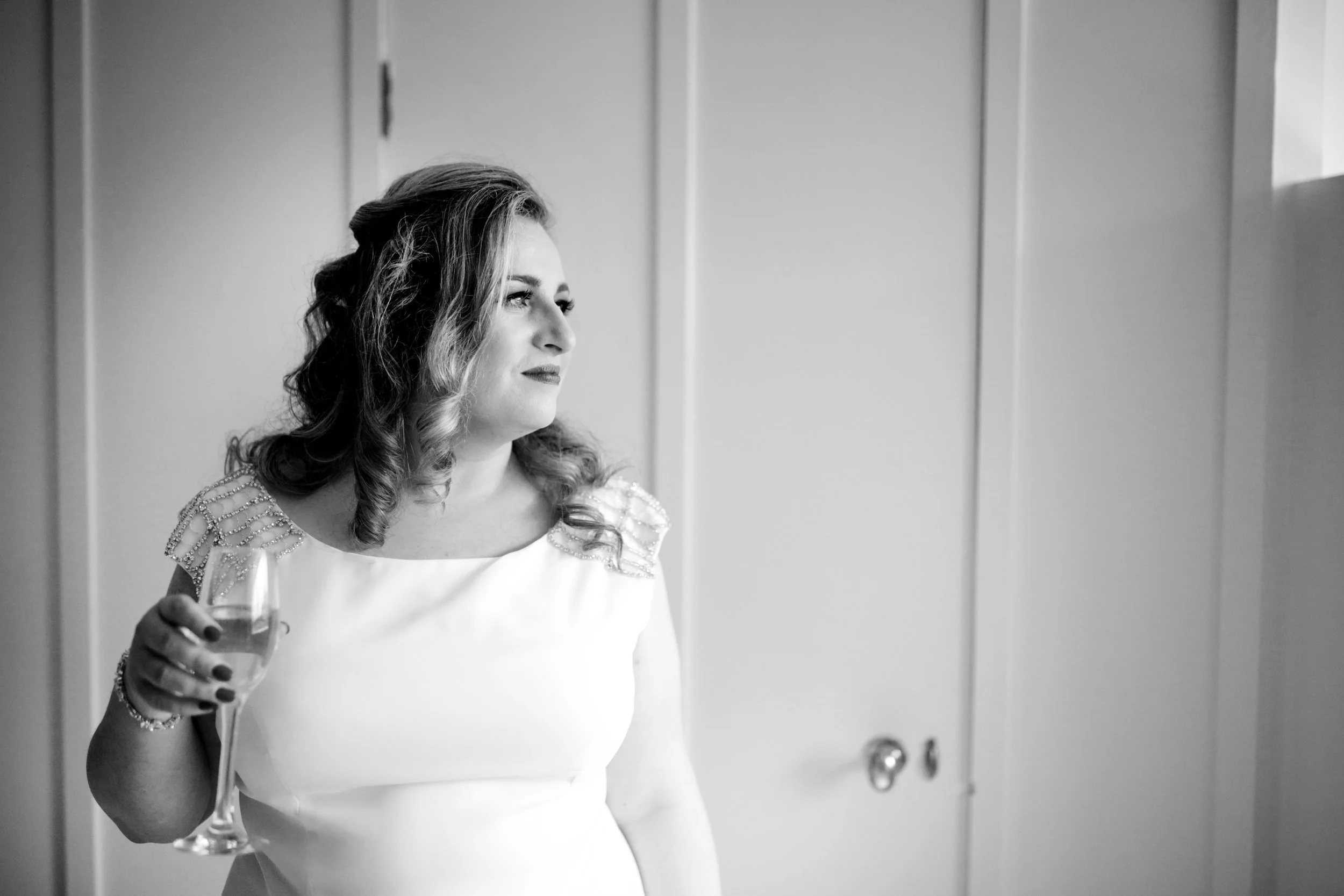 A woman with styled hair wearing a dress with embellished shoulders, holding a glass of wine, looking out a window in a bright room.