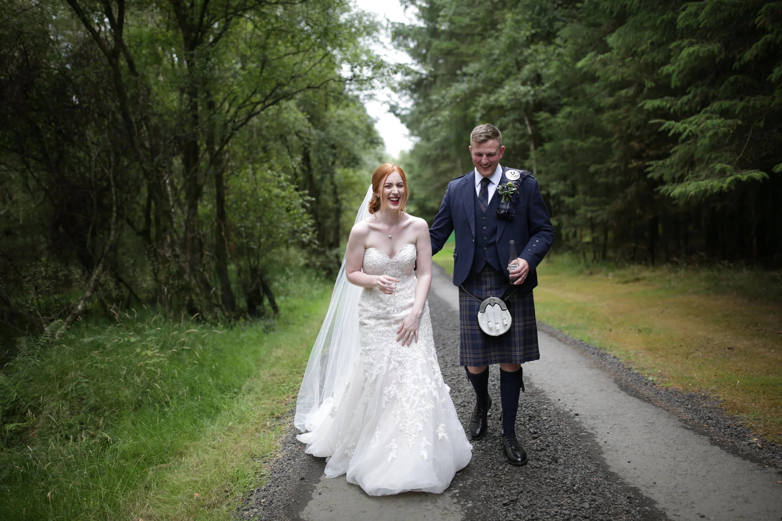 A bride and groom walking together on a forest trail, both smiling and happy, during their wedding day at Eden Leisure village, wedding venue Scotland