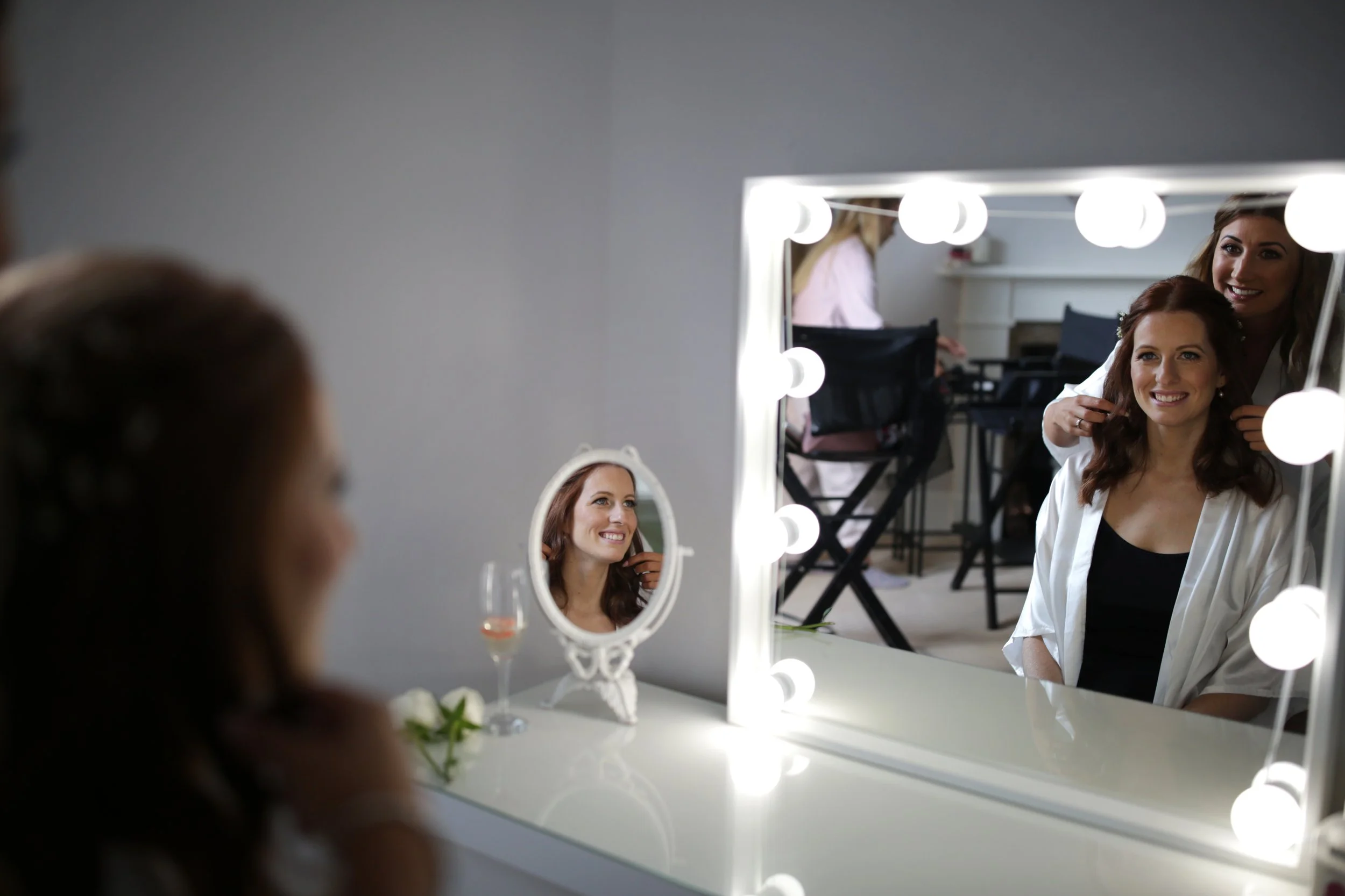 Bride smiling as a makeup artist arranges her hair in front of a mirror with light bulbs, with a smaller mirror and a glass of wine on the dressing table.