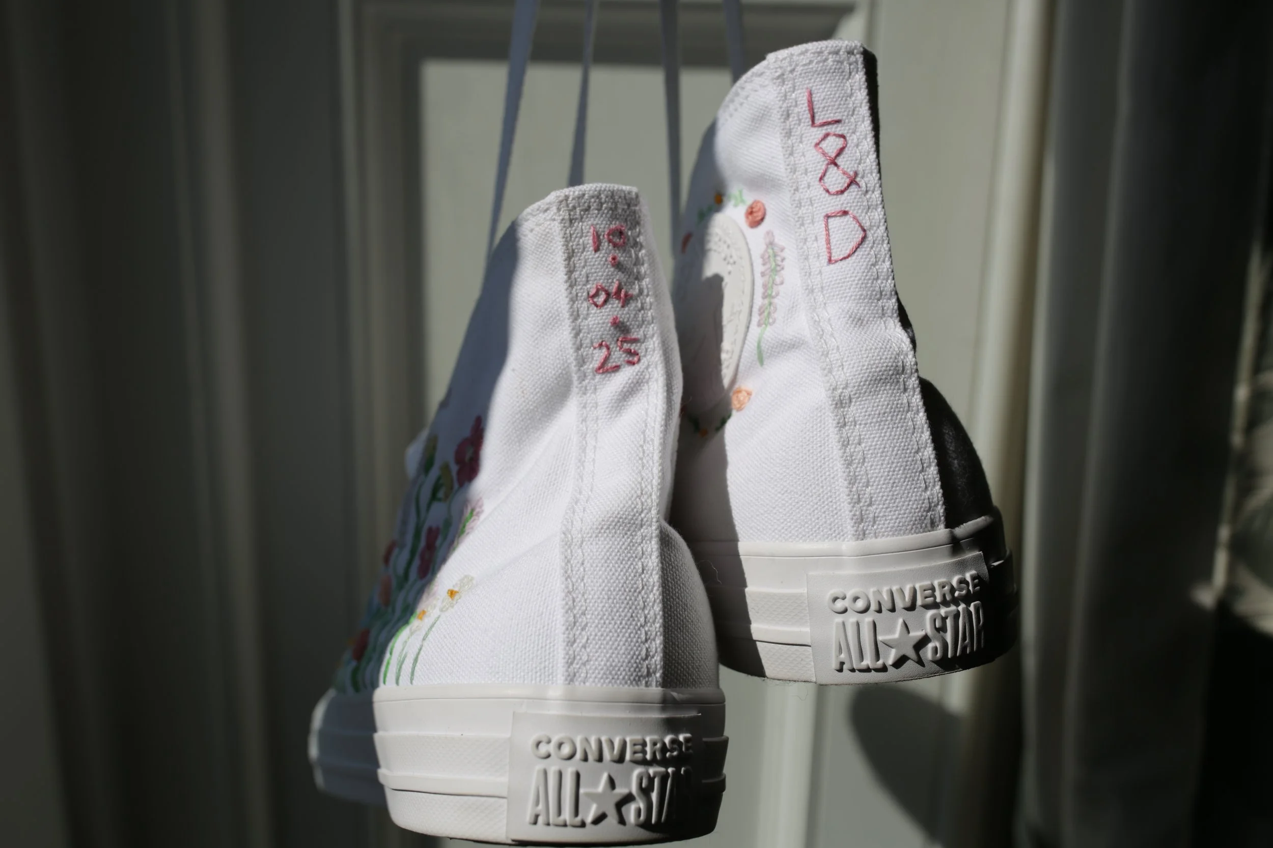 White high-top Converse All Star sneakers hanging indoors near a door, with colorful embroidery and red stitching on the heels.