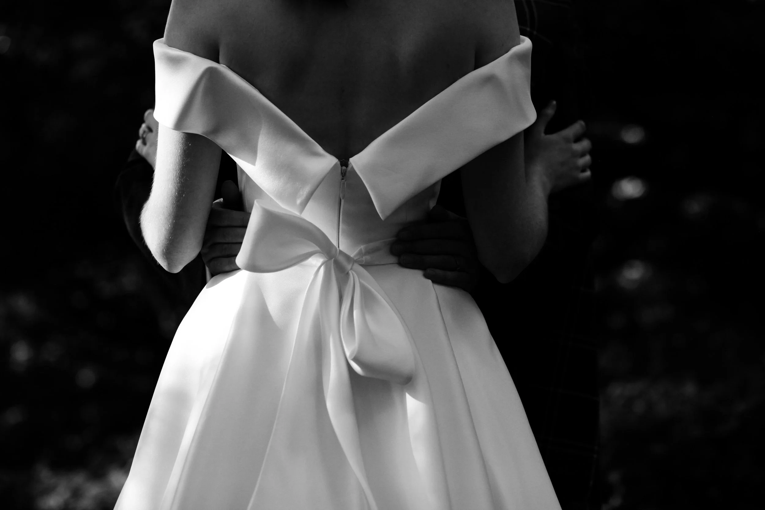 A person wearing an off-the-shoulder white dress with a large bow, being held by someone with hands around their waist, black and white photograph.