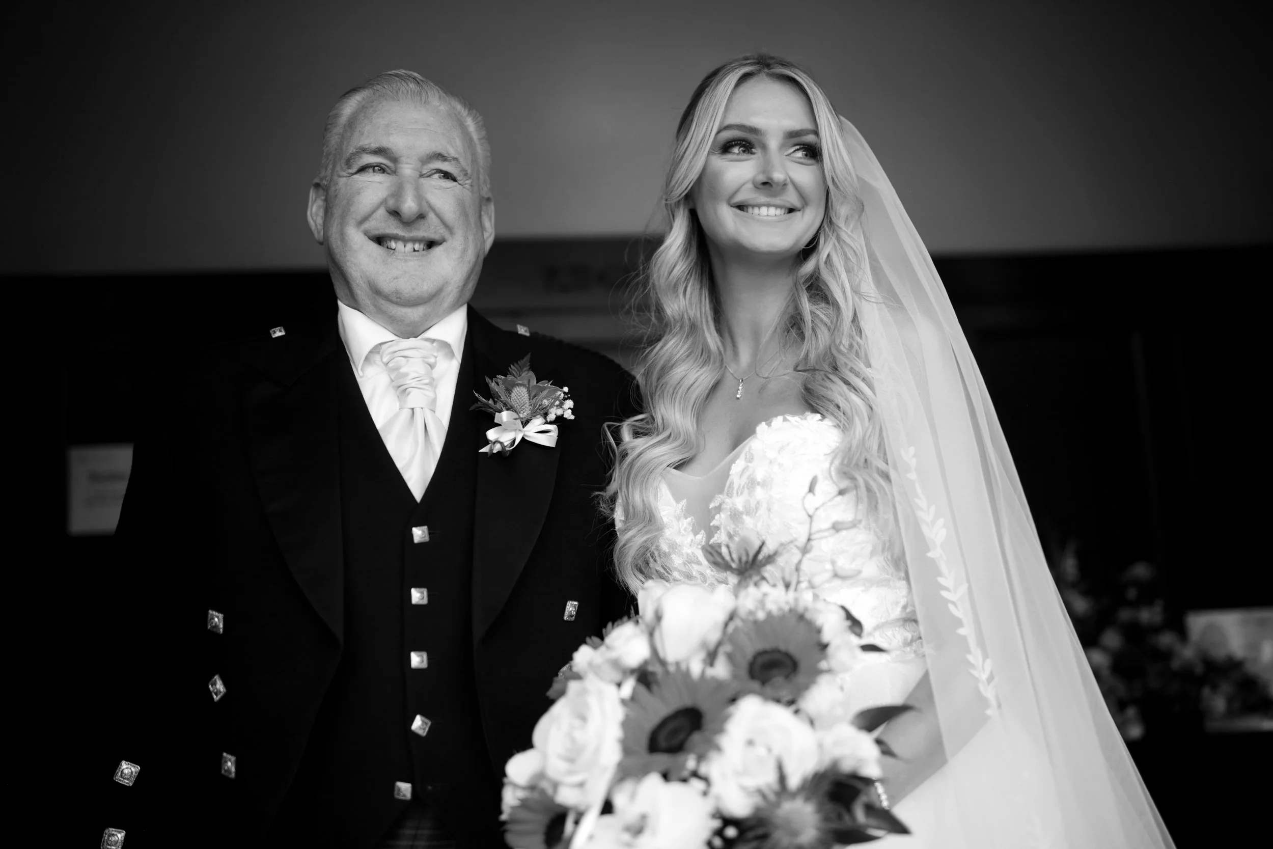 A groom with missing teeth and a bride with long wavy hair and a veil, smiling, standing together in a wedding setting.