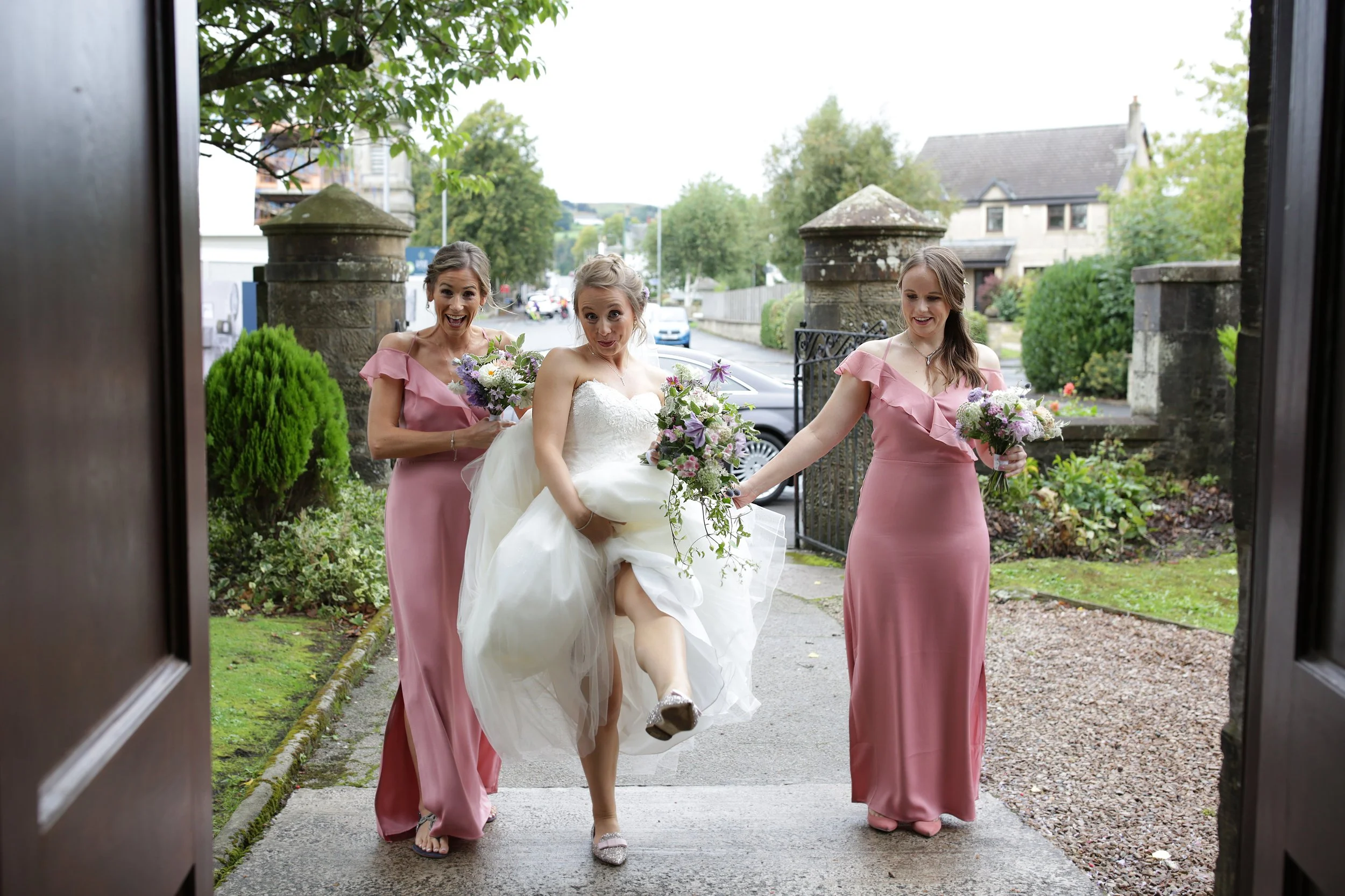 A bride in a white wedding dress being carried out by two bridesmaids in pink dresses, all holding bouquets of flowers, as they walk out of a building into a street with trees and houses.