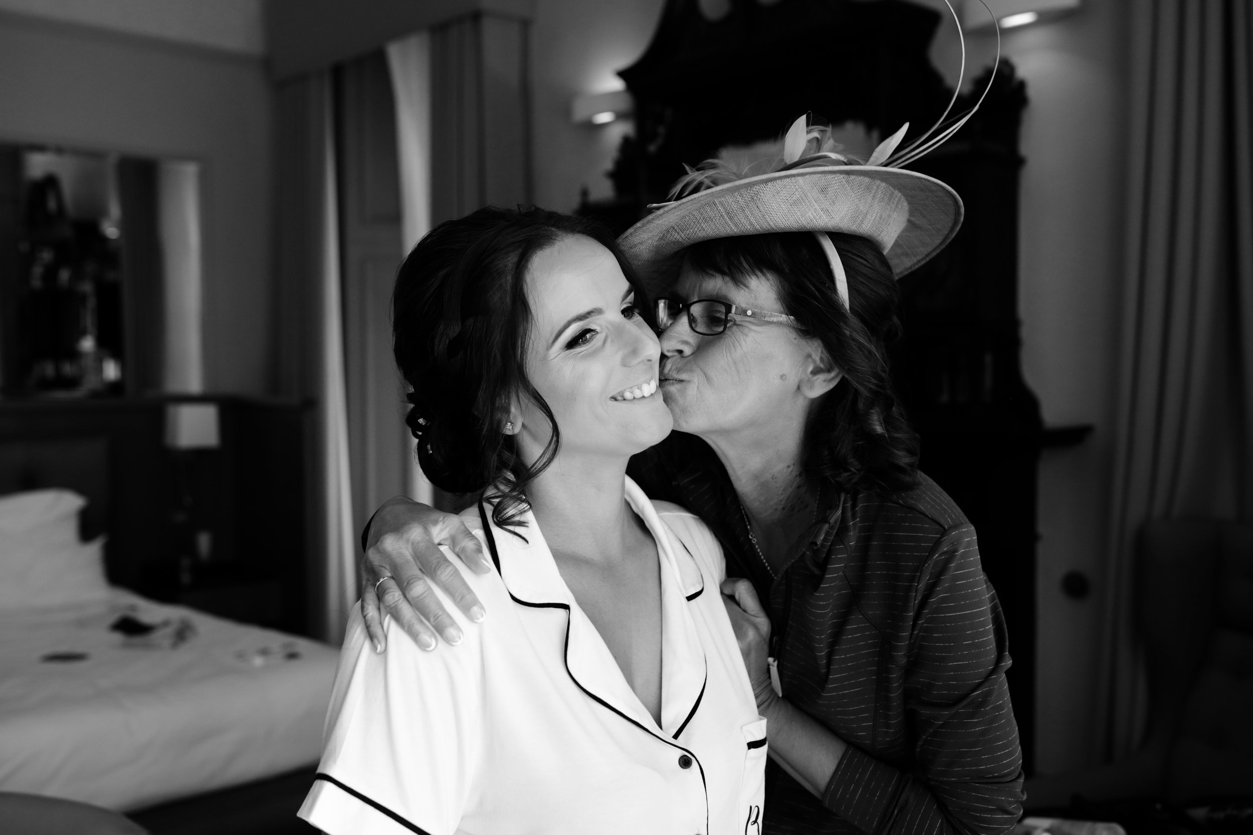 A woman with dark hair styled in curls, smiling as a woman wearing glasses and a decorative hat kisses her cheek in an indoor room with curtains and furniture in the background.
