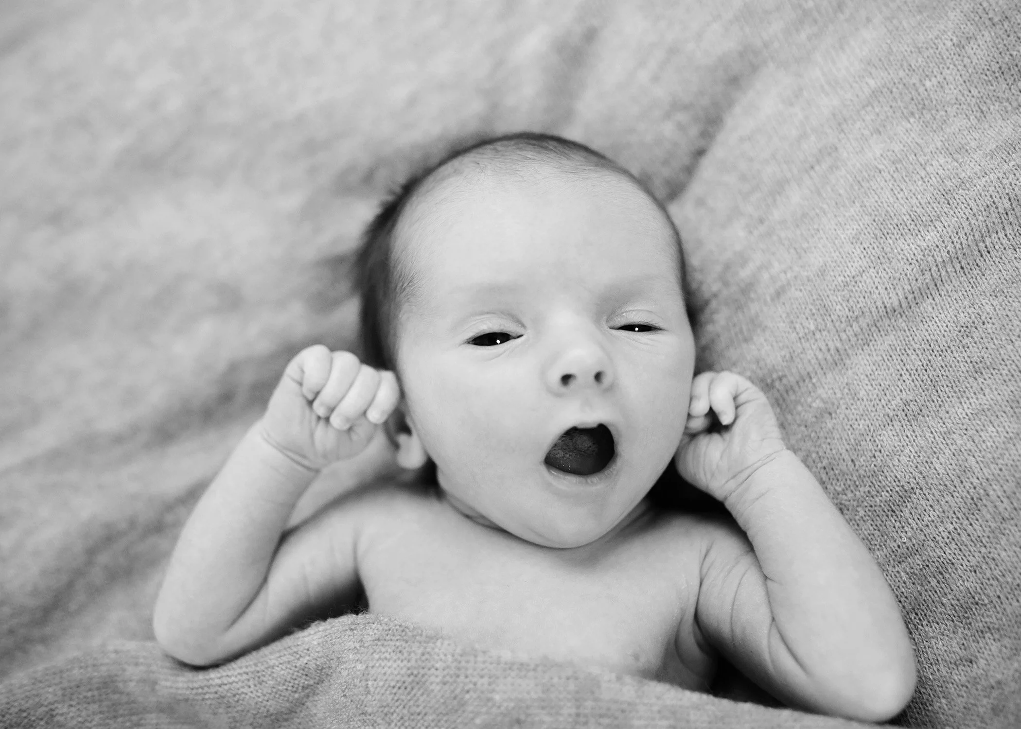 Black and white photo of a newborn baby lying on a soft surface, with arms bent and hands near the head, eyes partially open and mouth slightly open.