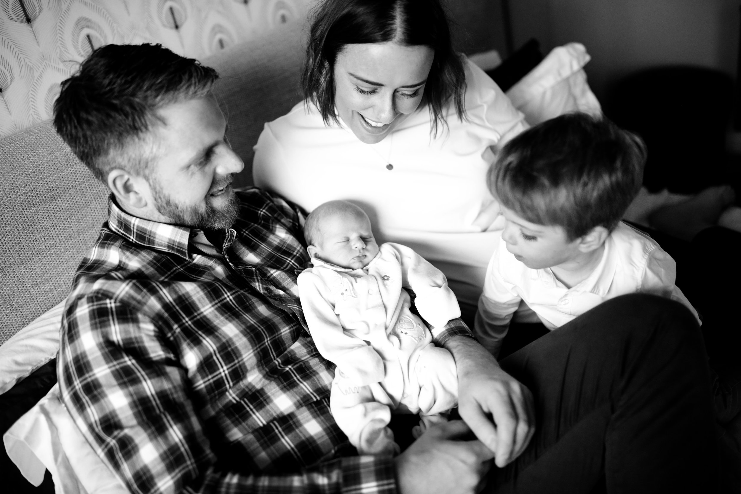 Black and white photo of a family with two children and a newborn baby, all gathered on a bed, sharing a joyful moment.