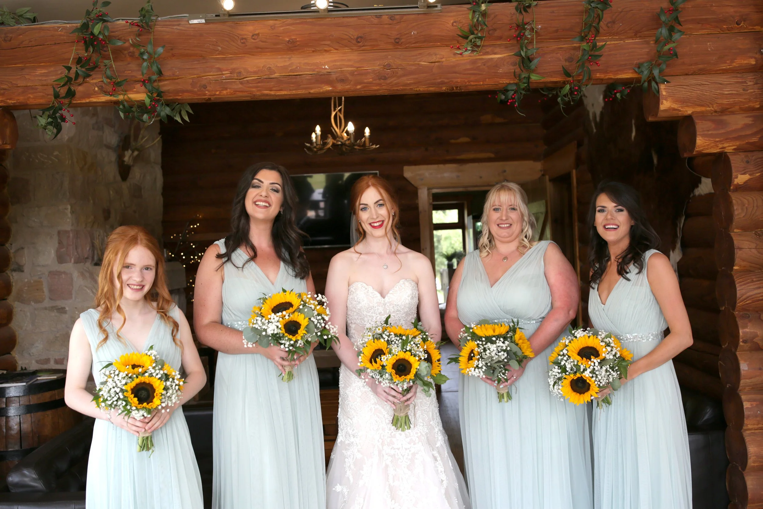 A bride and four bridesmaids standing indoors, each holding a bouquet of sunflowers and white flowers, in a rustic wooden room with a stone wall in the background.