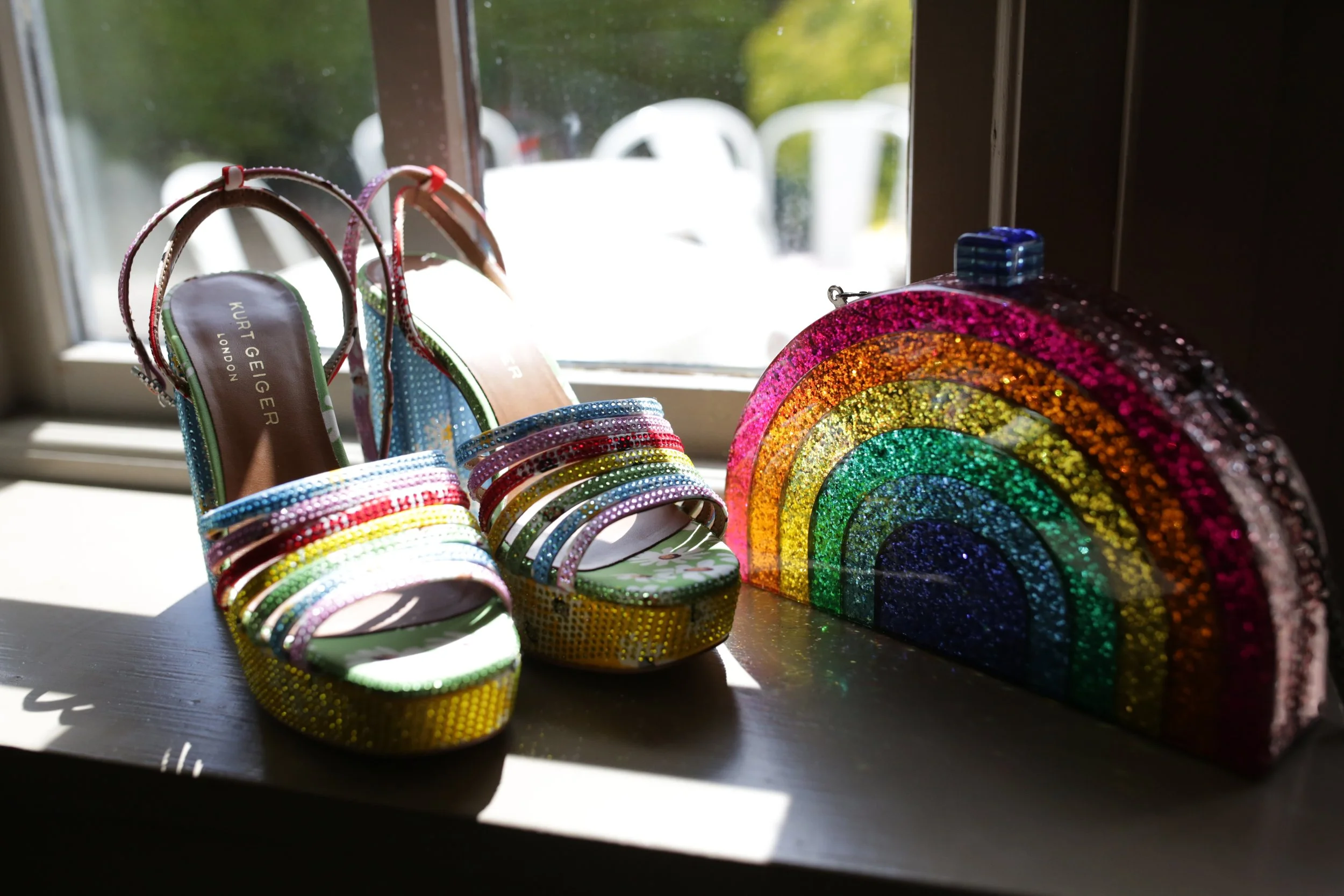 Rainbow-colored, glittery Kurt Geiger high-heeled sandals and a matching rainbow-shaped clutch purse on a windowsill, light shining through the window.