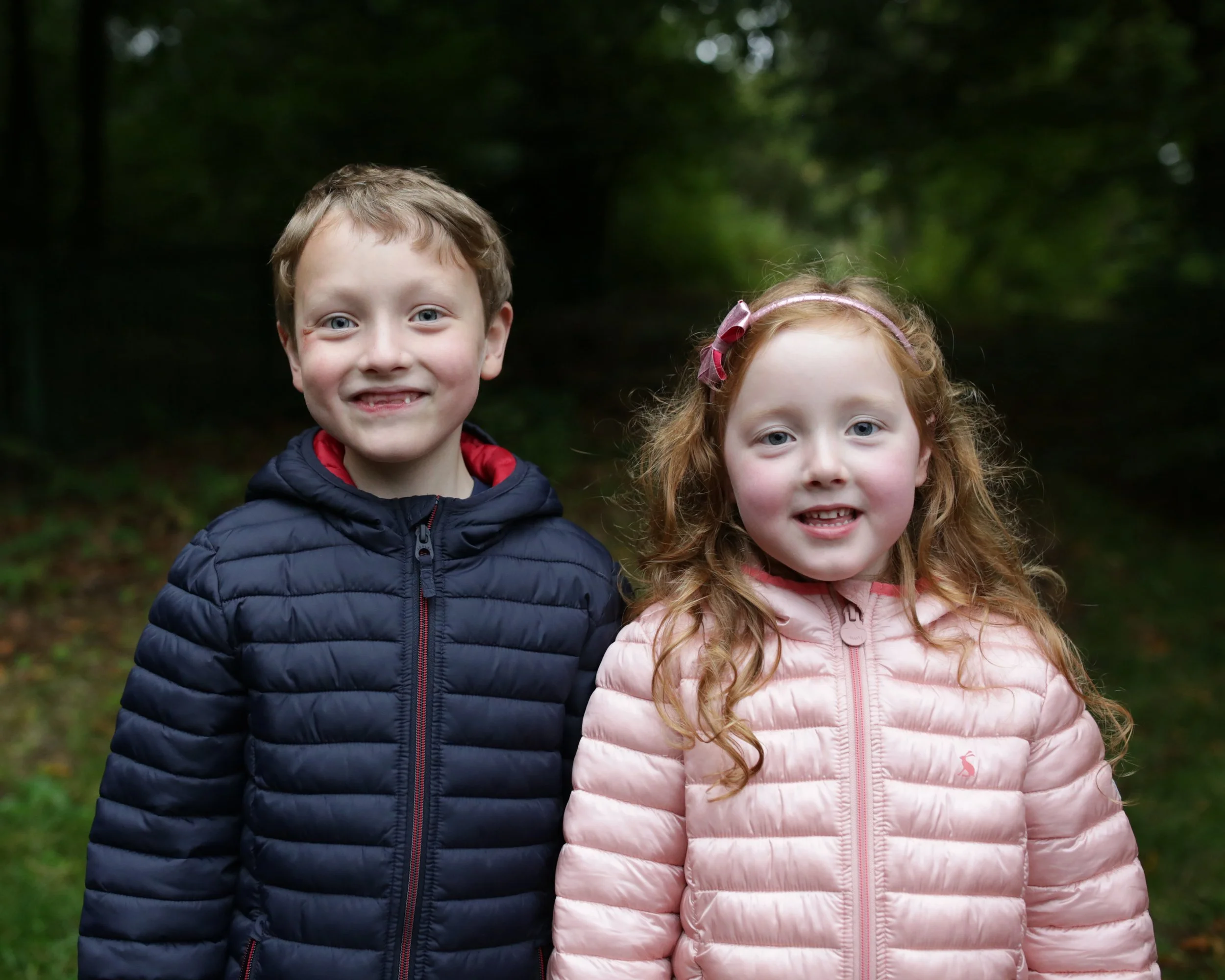 A young boy and girl standing outdoors in a wooded area, smiling at the camera, both wearing puffy jackets.