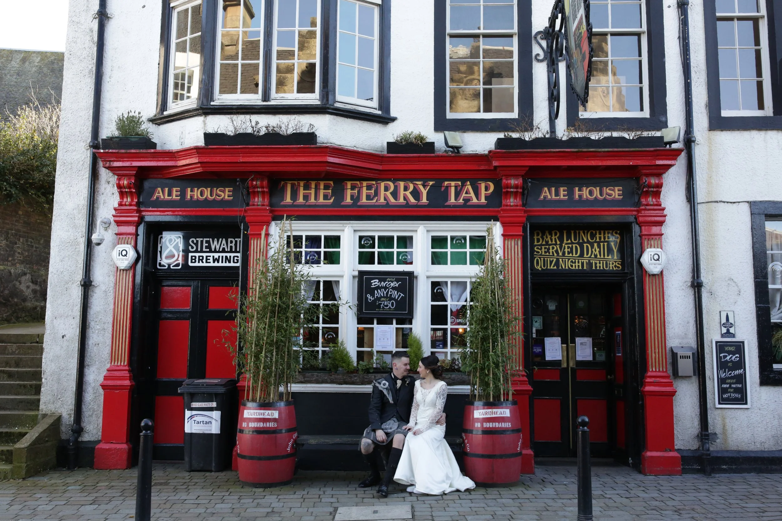 A couple in formal wedding attire sitting on a bench in front of a pub called 'The Ferry Tap'. The pub's exterior features red and black decor with signs advertising ale house, brewing, bar lunches, and quiz nights. The bride wears a white wedding dr