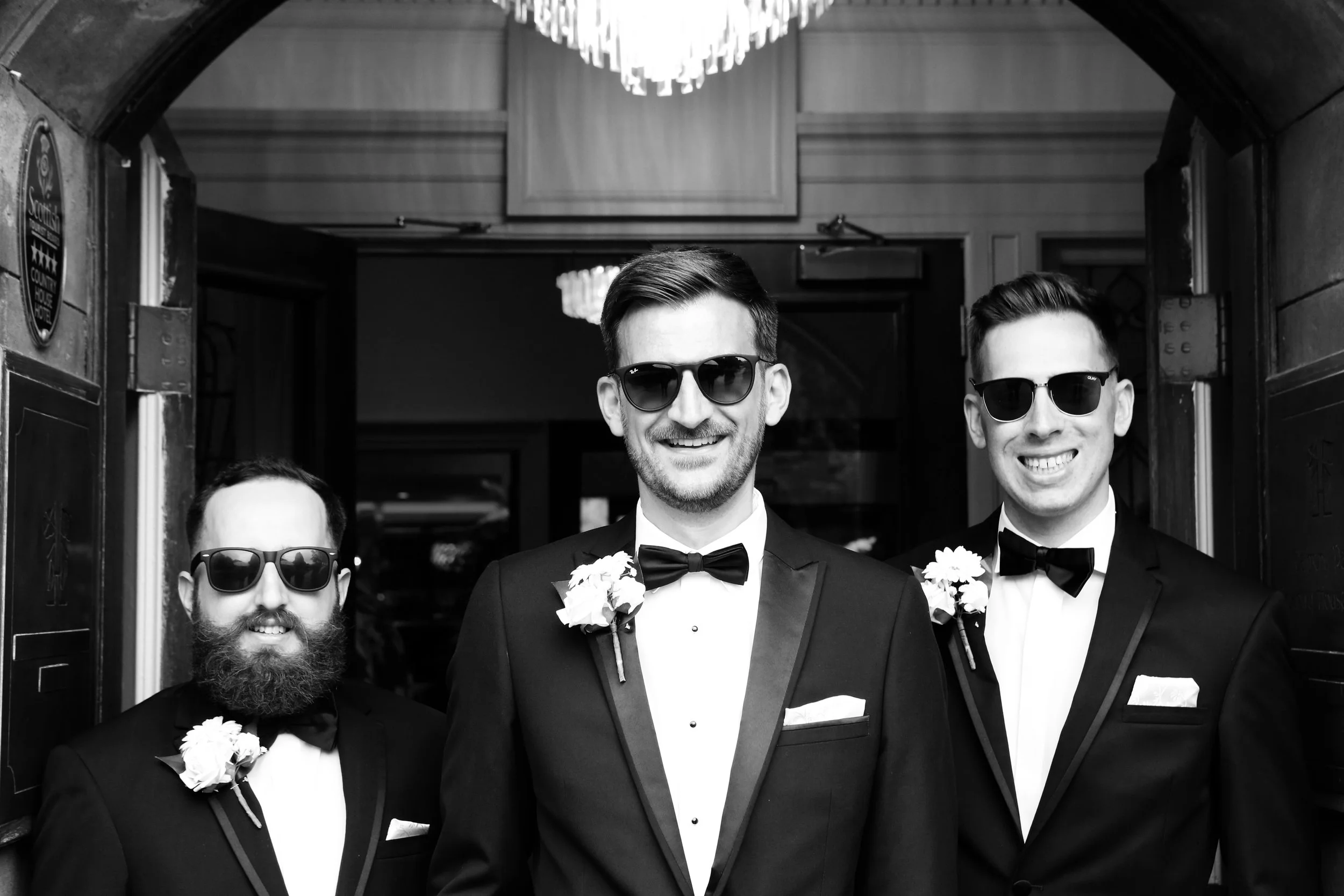 Three men in tuxedos and sunglasses posing for a photo at a formal event, with one man in the middle and two men on either side, each wearing a boutonniere on their tuxedo lapels.