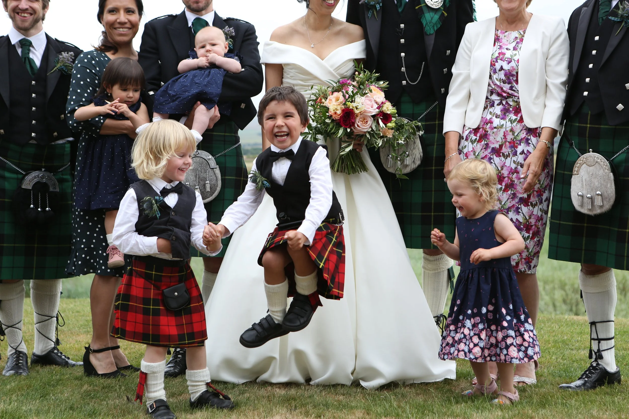 Group of people, including children and adults, at a wedding outdoors, with a woman in a white wedding dress holding a bouquet, children in kilts and dresses, and the children in the foreground appear happy and playful.