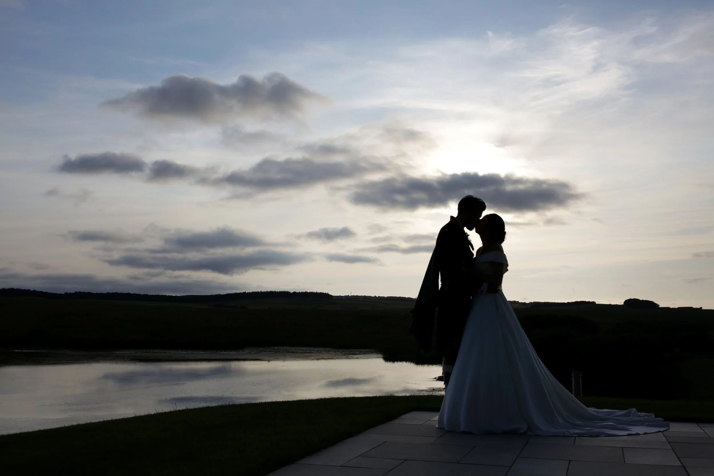 Silhouette of a couple in wedding attire standing close together by a body of water during sunset.