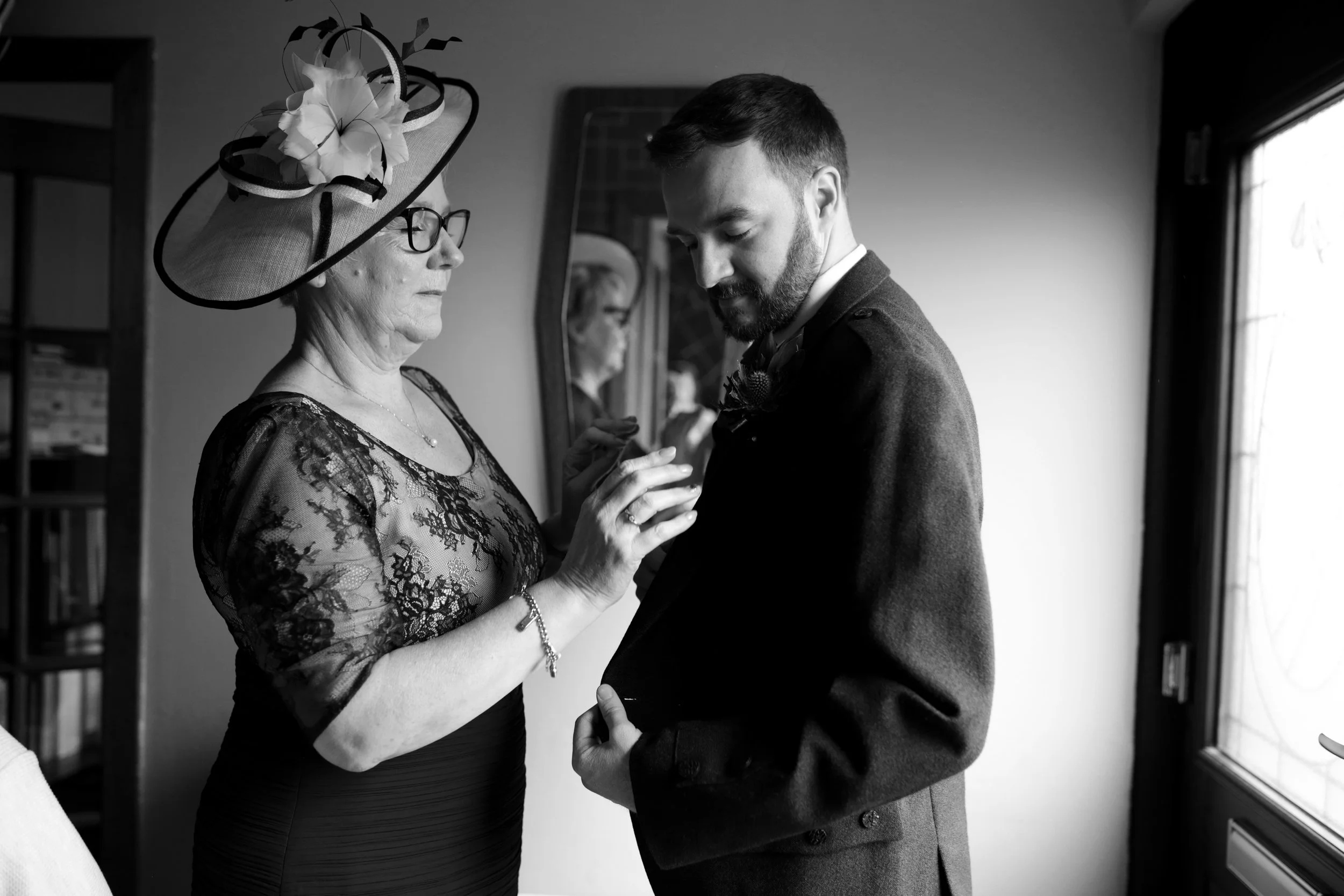 An elderly woman placing a pin on a groom's lapel during a wedding preparation, both looking down at the boutonniere, with a mirror reflecting the woman’s face.