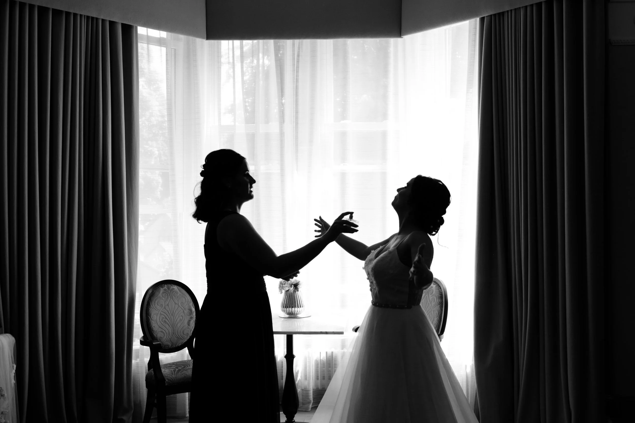 Silhouette of two women, one in a wedding dress, in a room with large window and curtains.