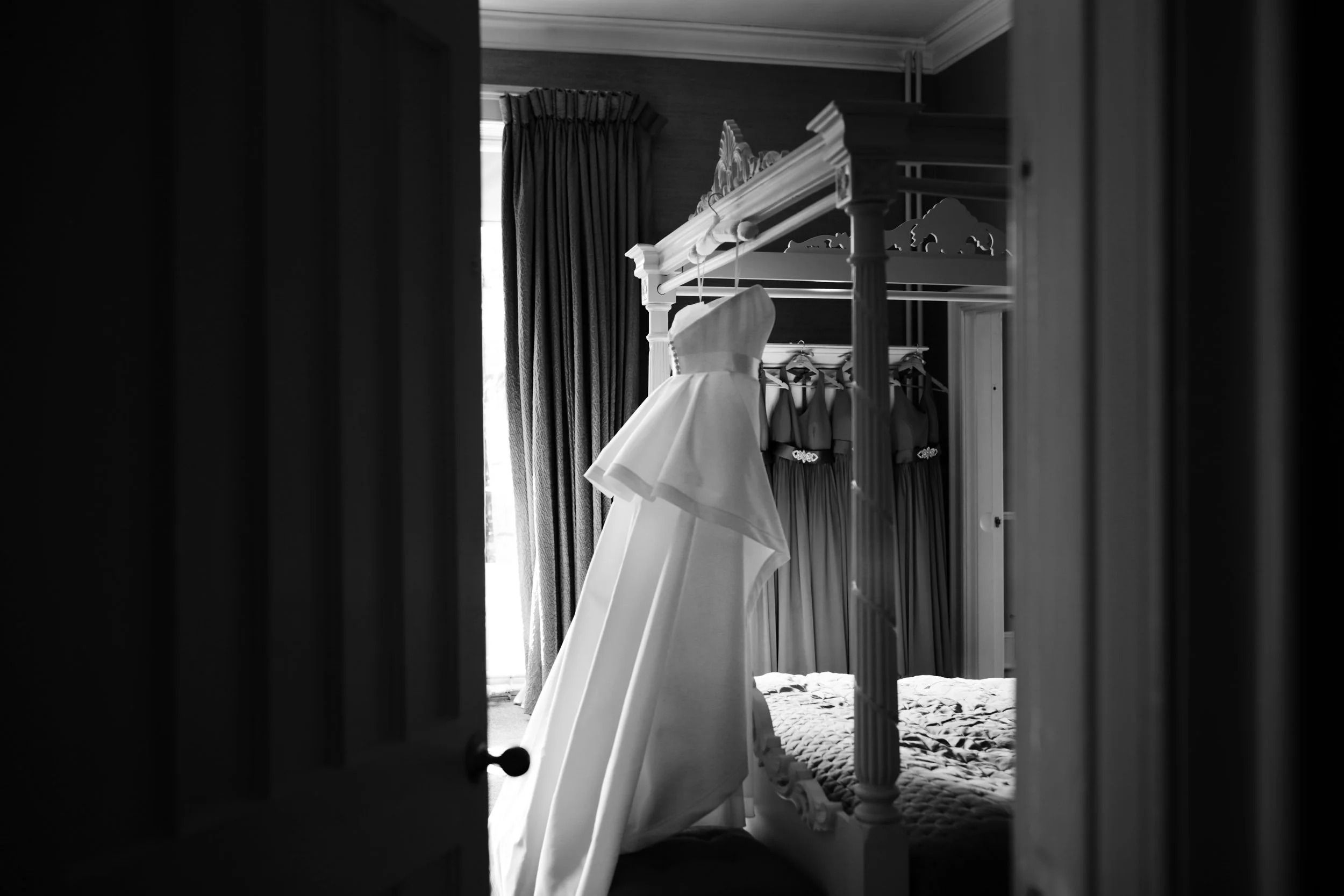 A wedding dress hanging in a bedroom with natural light coming through the window, seen through a partially open door.