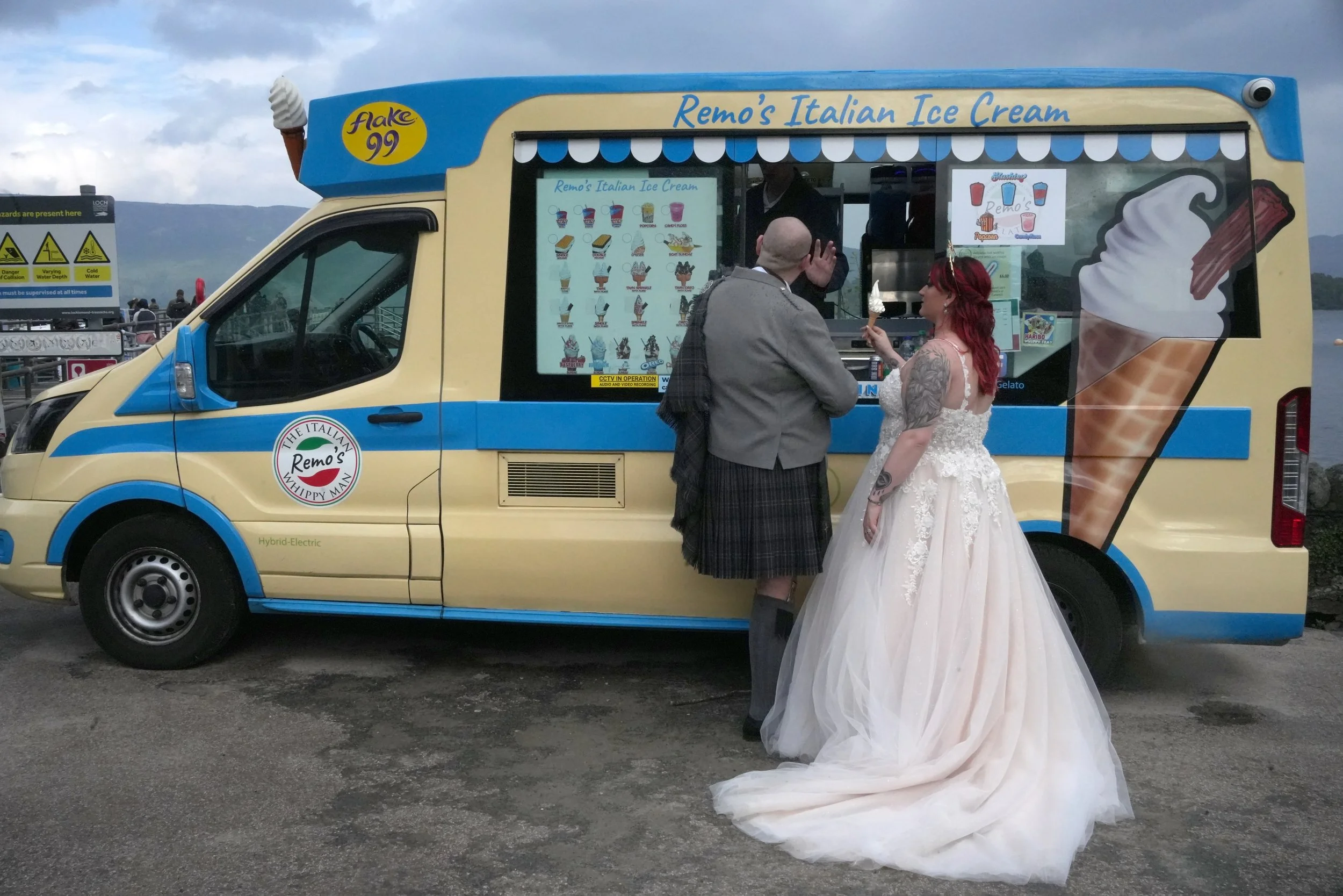 A wedding couple is standing in front of a colorful ice cream truck, with the bride in a white wedding gown and the groom in a kilt and jacket. Loch Lomond Arms, Wedding photography Scotland, Wedding Venue Scotland. 