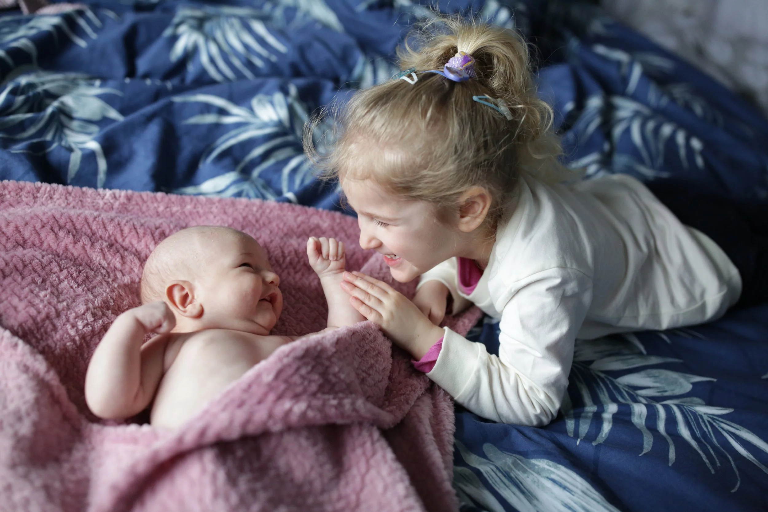 A young girl and a baby sharing a joyful moment on a bed, with the girl leaning over the baby and smiling as they touch hands.