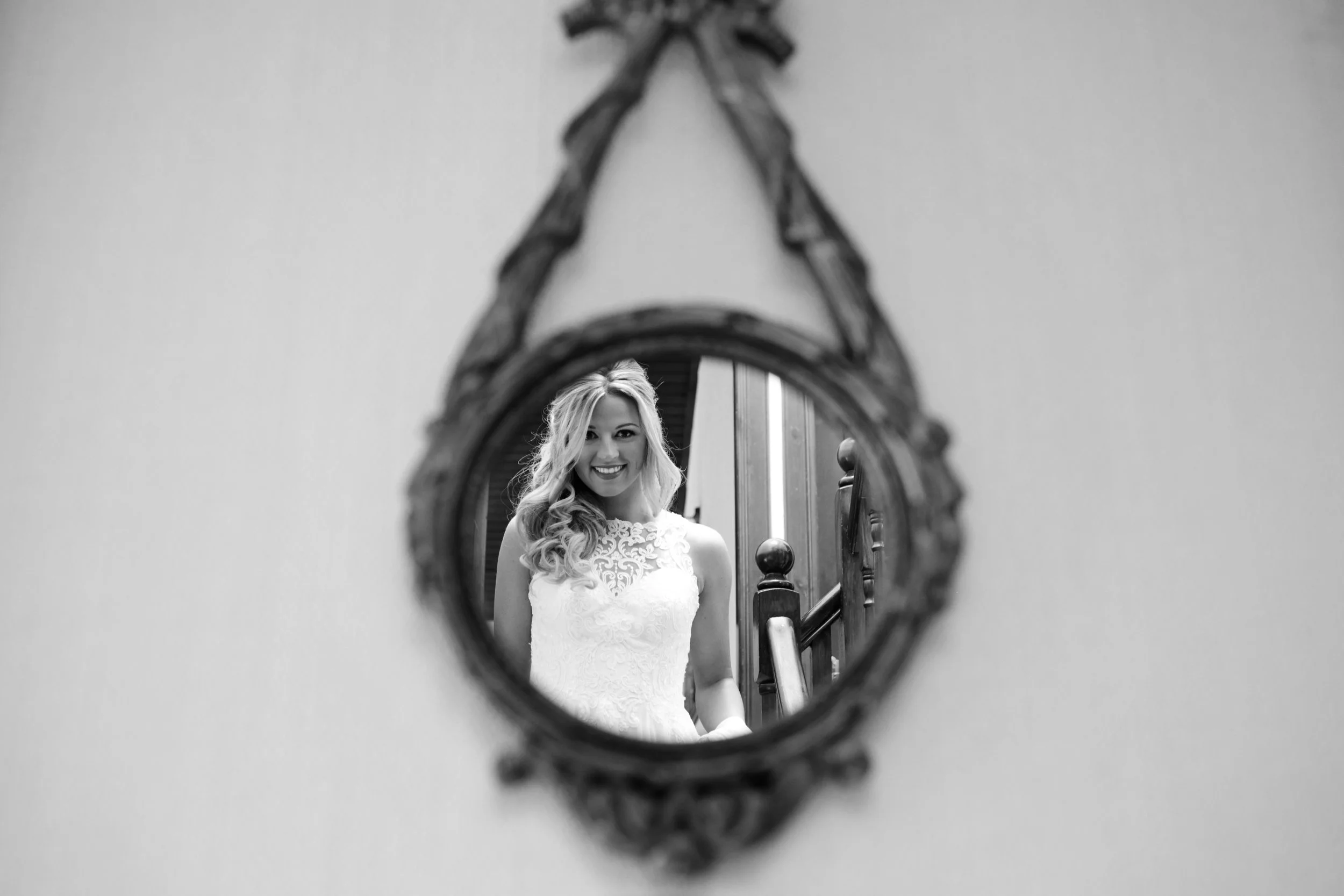 A woman in a white lace dress is smiling as her reflection is seen in an ornate oval mirror on the wall.