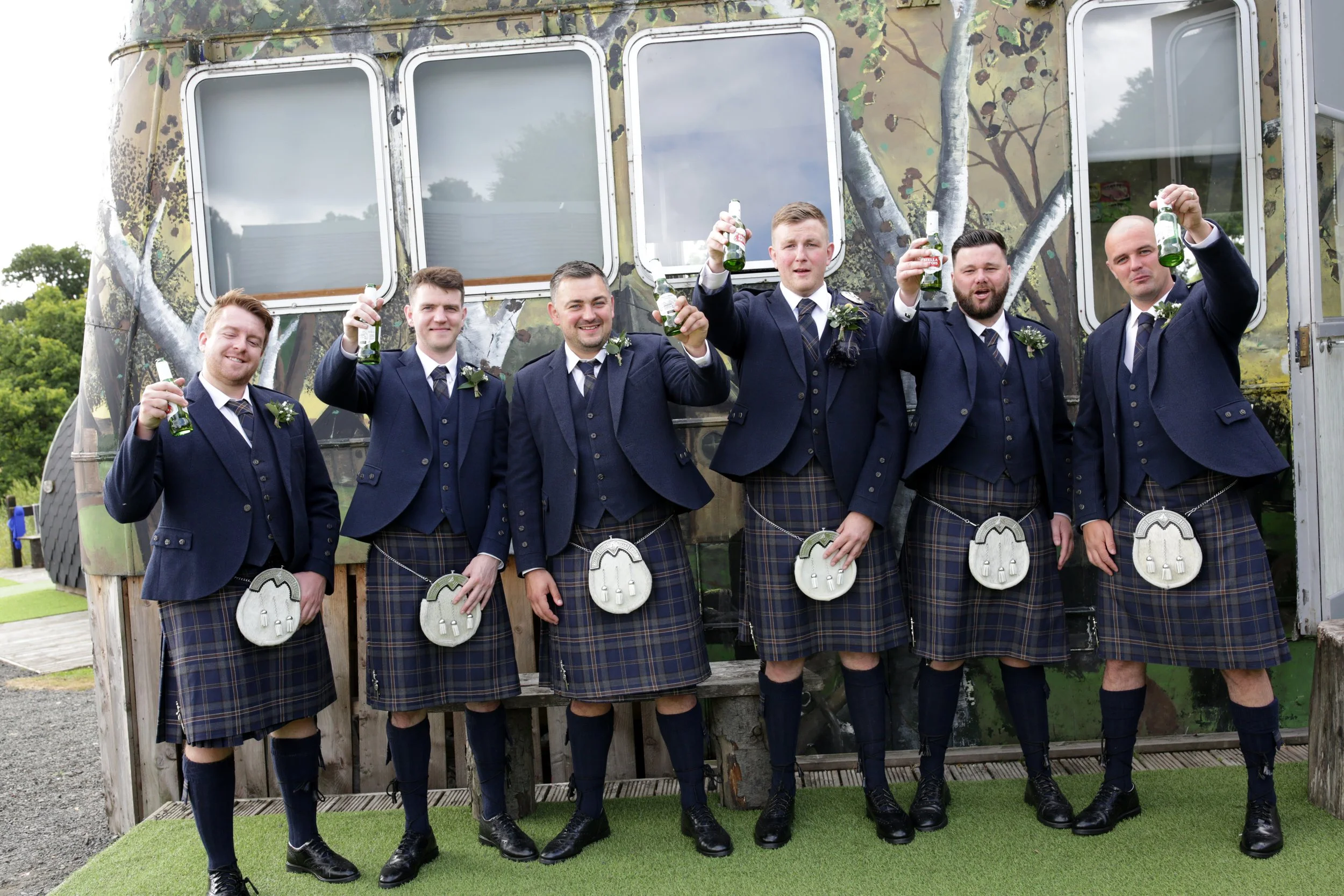 Six men dressed in traditional Scottish kilts and suits, standing outdoors in front of a painted trailer, raising beer bottles in celebration.