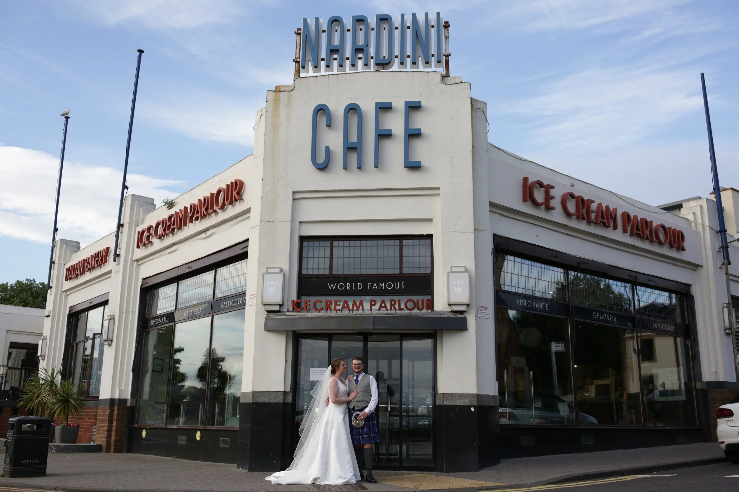 A bride and groom standing in front of Nardini Cafe, an ice cream parlor, with large glass windows and signage for Italian bakery, ice cream, and parlor, on a city street.
