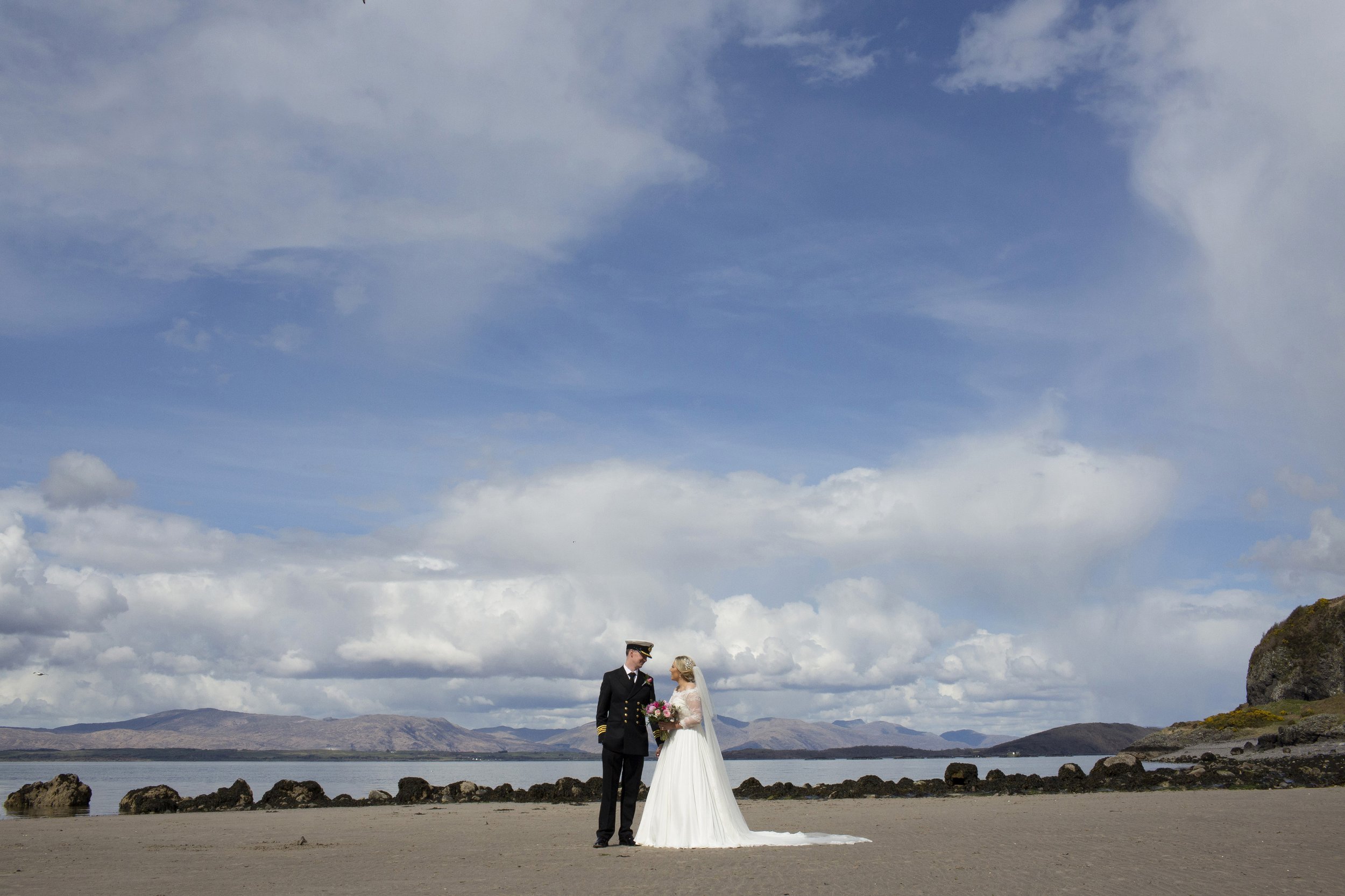 Bride and groom standing on a sandy beach, holding hands, with mountains, water, and partly cloudy sky in the background.