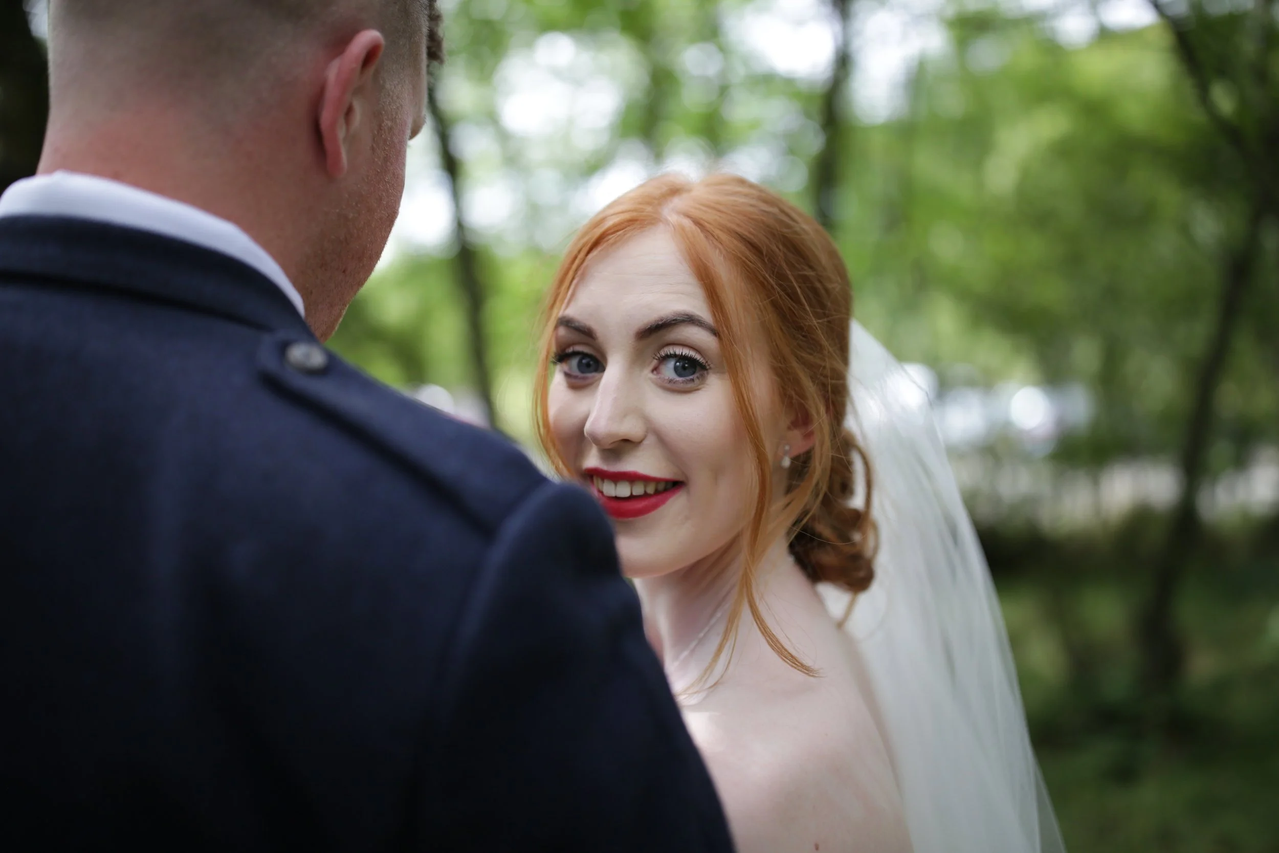 A bride with red hair, wearing a white wedding dress and veil, smiling at the camera while standing outdoors with a groom in a dark suit beside her, in a green, wooded area.