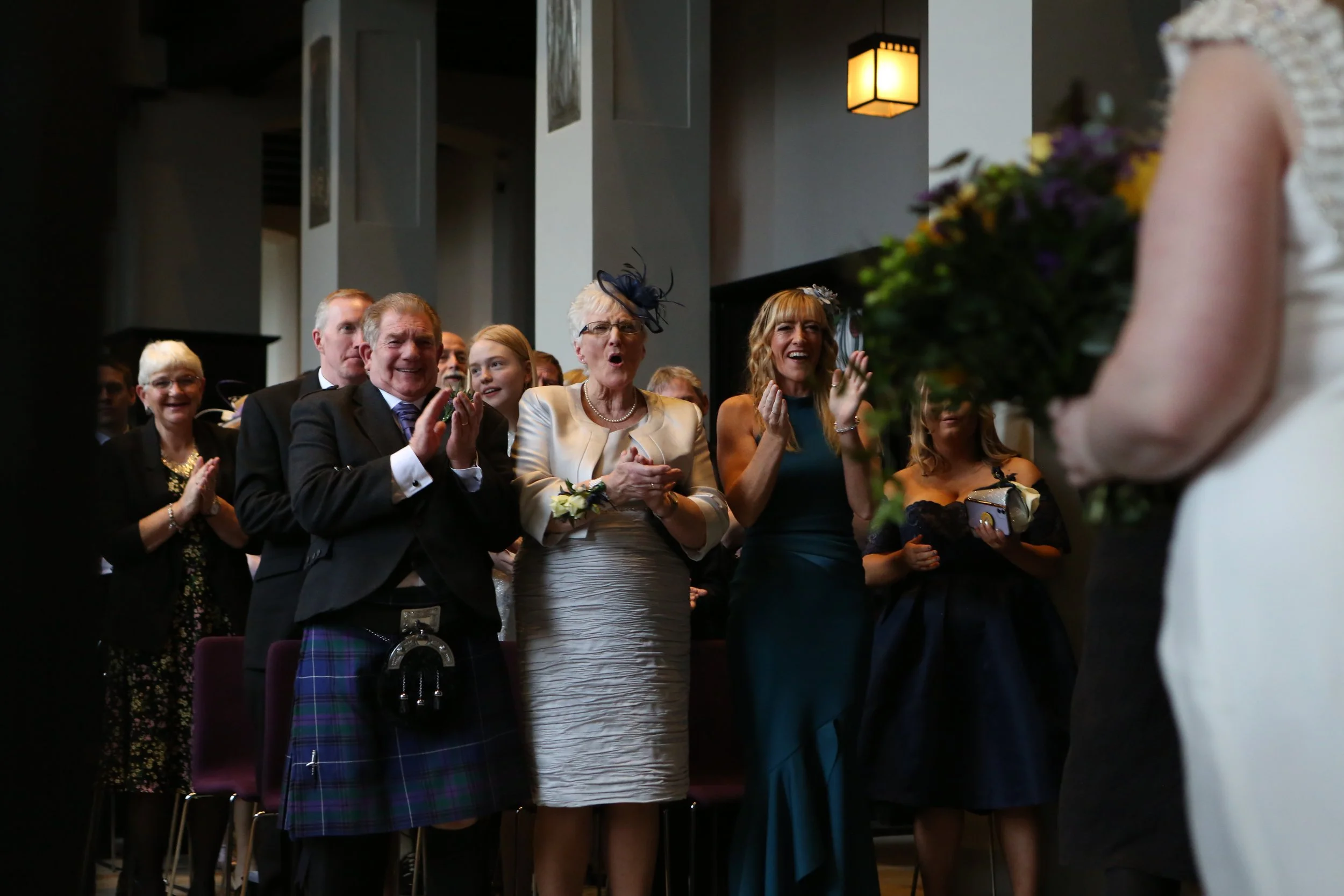 People at a wedding ceremony reacting with joy and surprise as they look at a bride holding a bouquet.