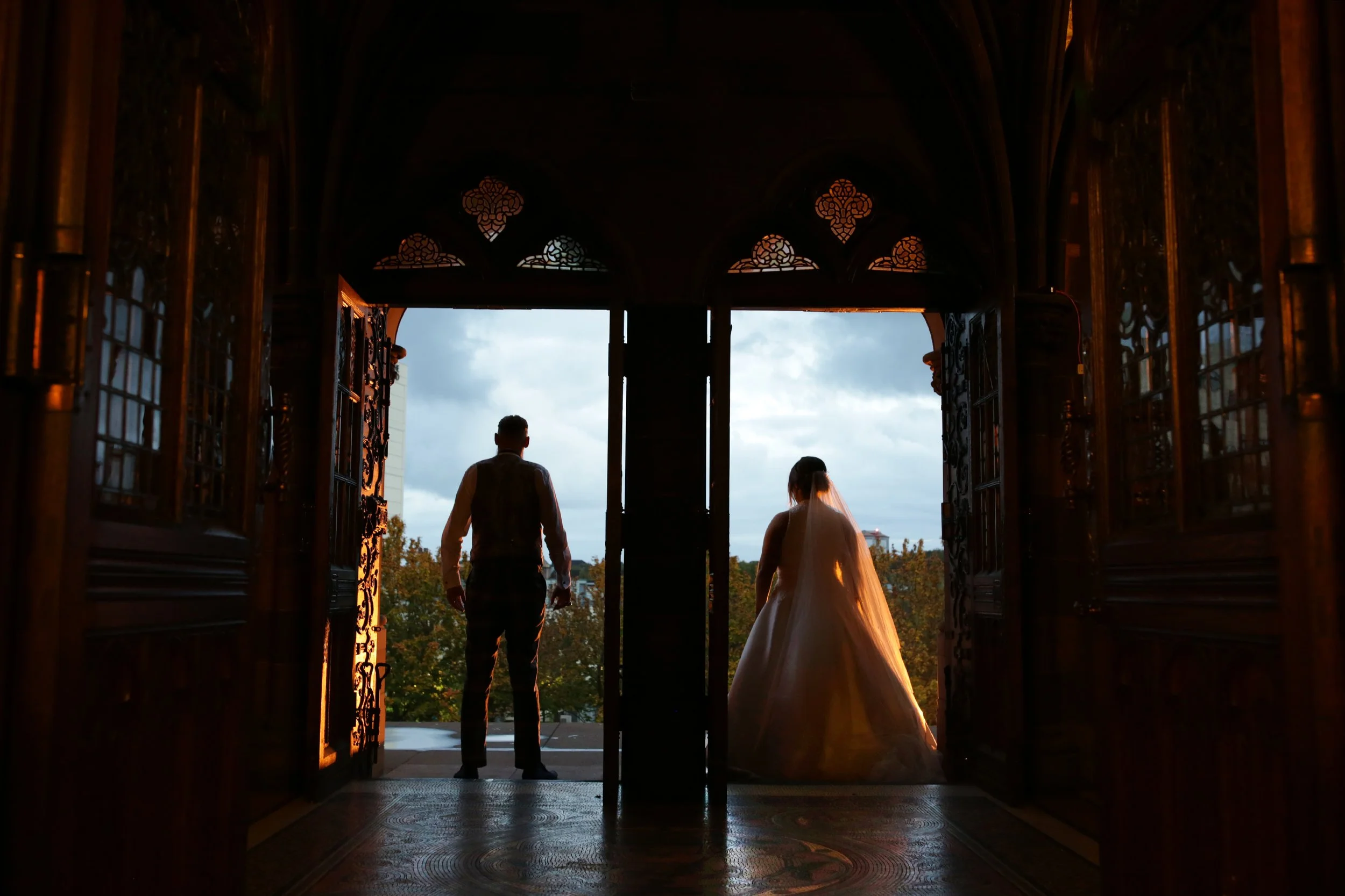 A bride and groom standing on a balcony at sunset, silhouetted against a cloudy sky, framed by ornate wooden doors.