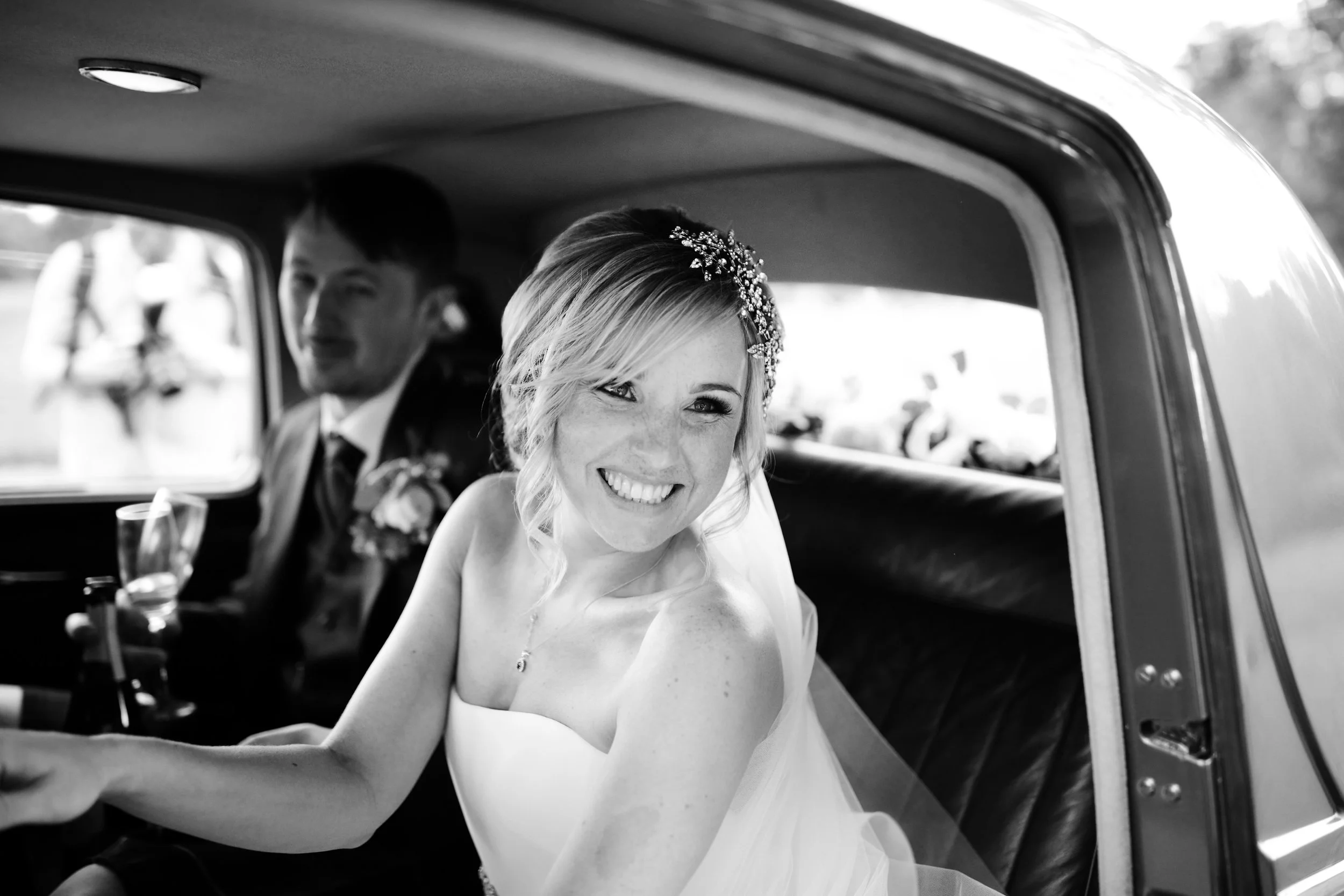 Smiling bride with a floral headpiece sitting in a vintage car, with groom blurred in background.