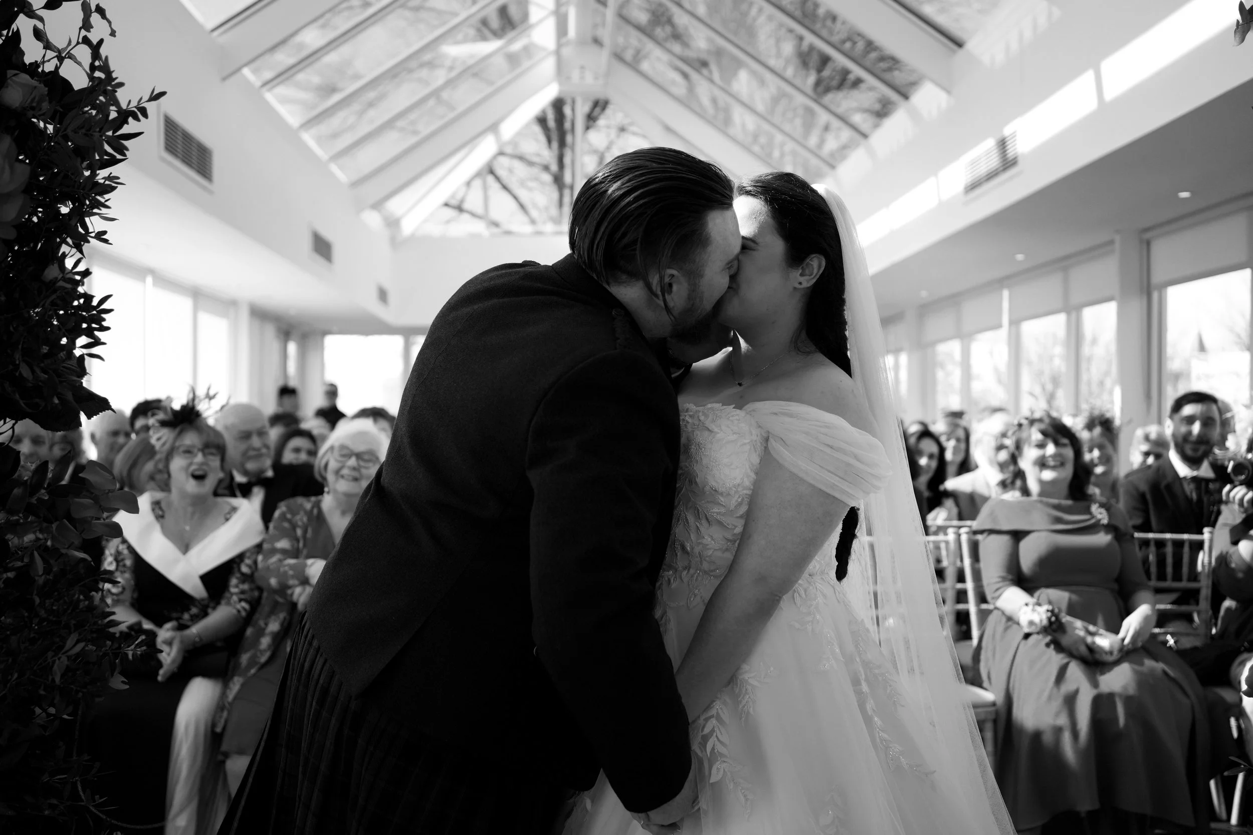 A bride and groom kiss during their wedding ceremony inside a bright, glass-ceiling venue, with guests seated and smiling in the background.